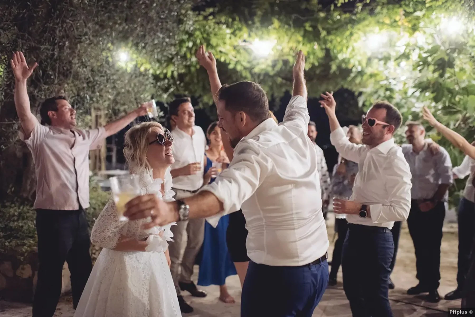 Festa di matrimonio all'aperto con persone che ballano e si divertono sotto le luci, alcune indossano occhiali divertenti.