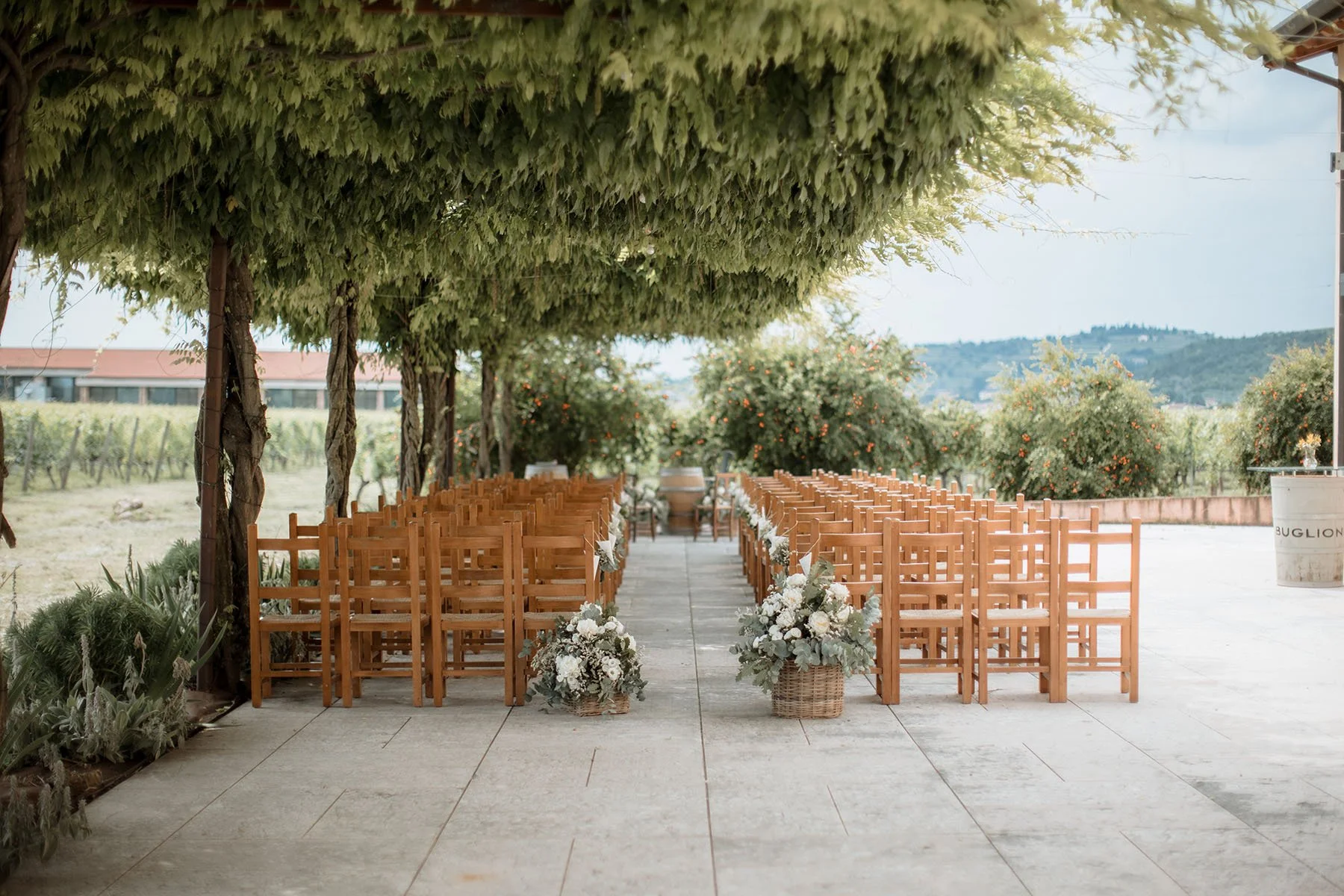 Cerimonia all'aperto con sedie di legno e decorazioni floreali in un'ambientazione naturale con alberi e panorami collinari.