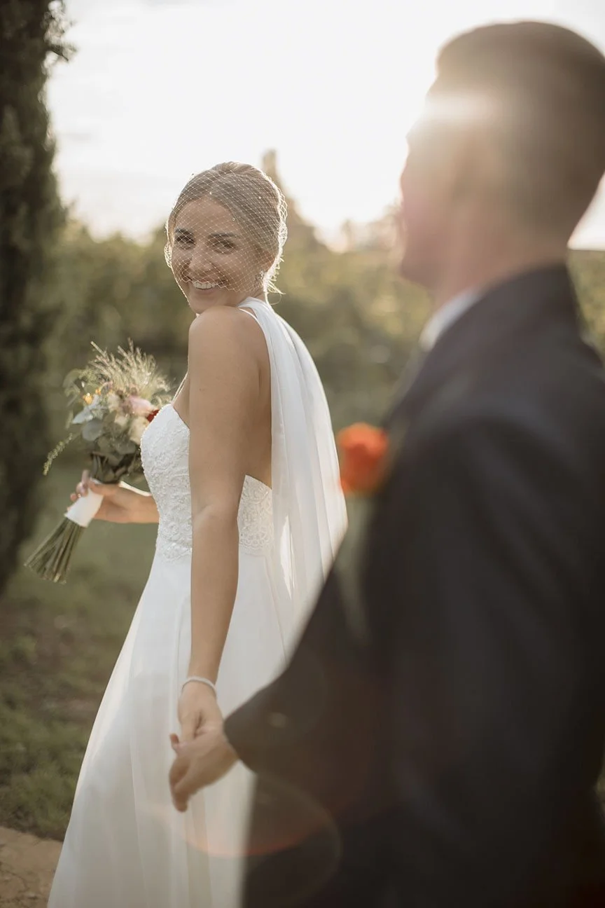 Una coppia di sposi si tiene per mano all'aperto al tramonto, la sposa sorride mentre guarda verso l'obiettivo e indossa un abito da sposa bianco con velo, la figura dello sposo è sfocata.