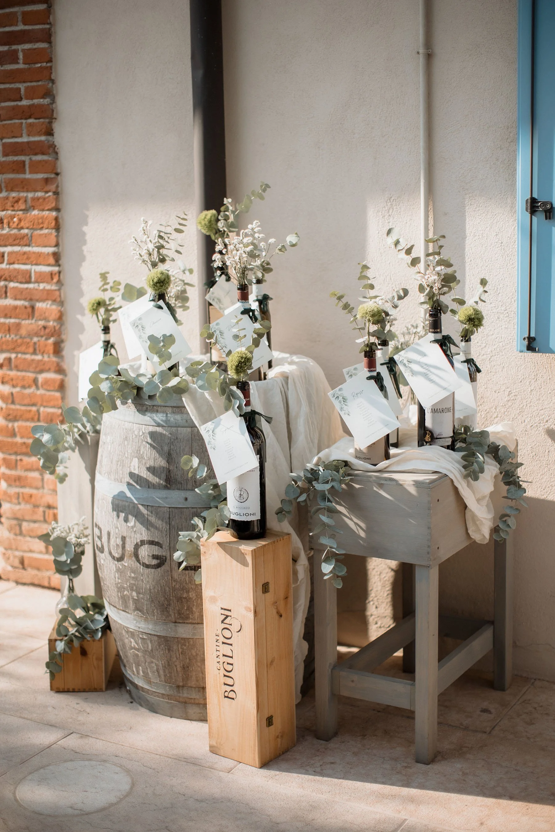 Bottiglie di vino decorativo con foglie di eucalyptus e note di ringraziamento, posizionate su un tino di legno e un tavolino di legno, in un ambiente con pareti di pietra e intonaco con luce naturale.