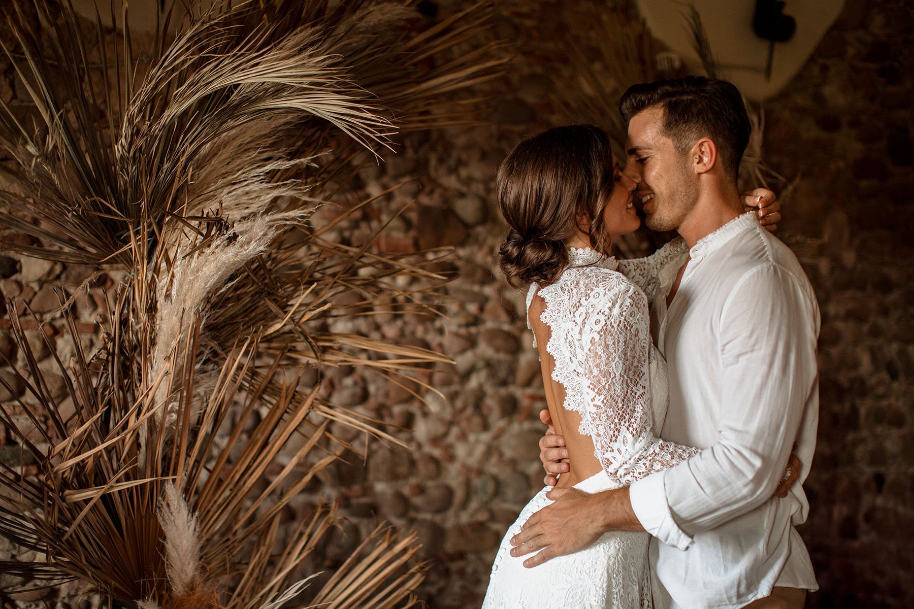 Una coppia innamorata in un abbraccio, con il volto molto vicino, sorridenti, davanti a uno sfondo di pietre e grandi piante di pampas grass.