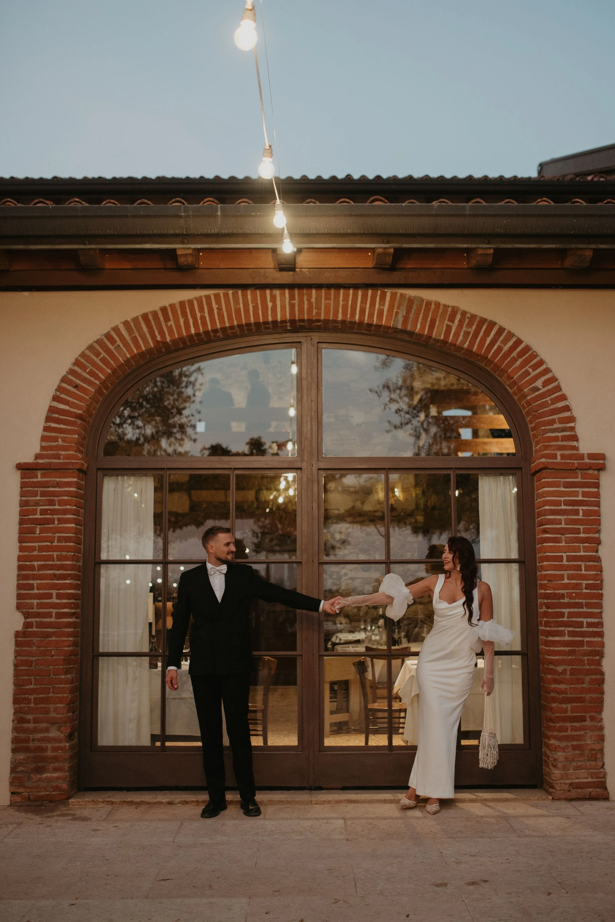 Coppia di sposi che si tiene per mano davanti a una grande porta di vetro con cornice in legno e mattoni rossi, all'aperto in un'atmosfera serale.
