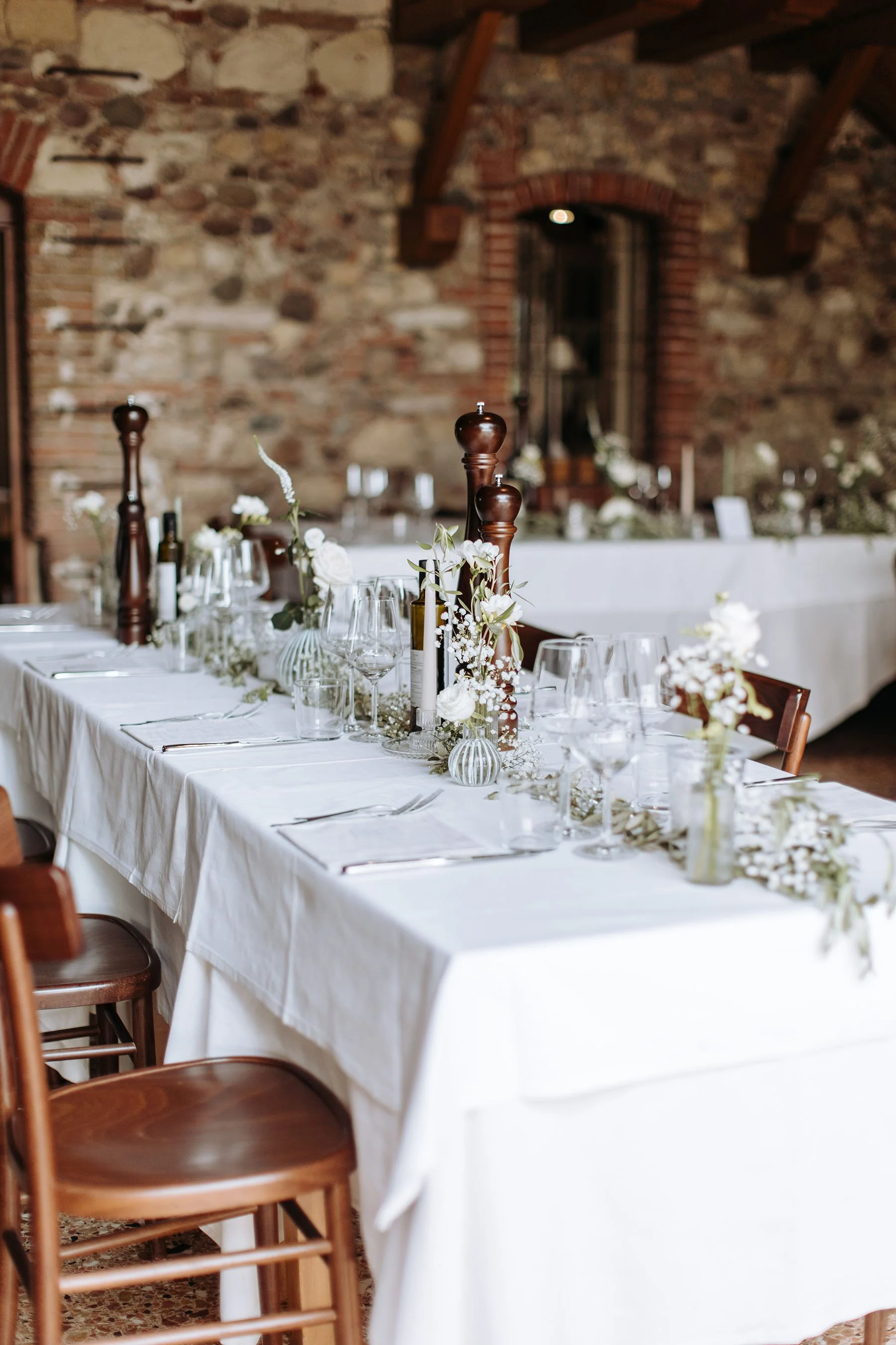 Tavola imbandita decorata con fiori bianchi e candele, all'interno di una stanza con pareti in pietra e soffitto in legno