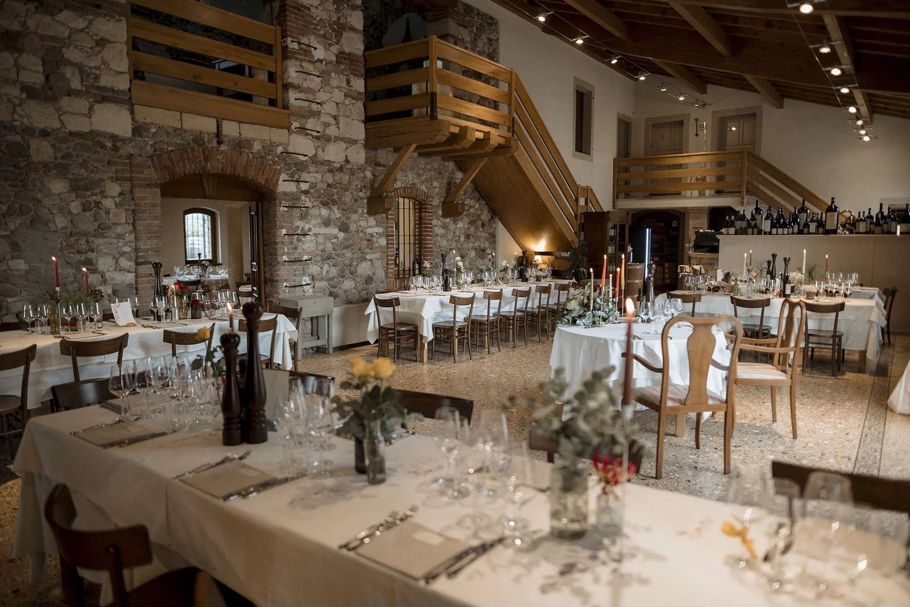 Sala per un evento con tavoli imbanditi, candele e decorazioni floreali in un ambiente rustico con pareti di pietra e soffitto in legno.