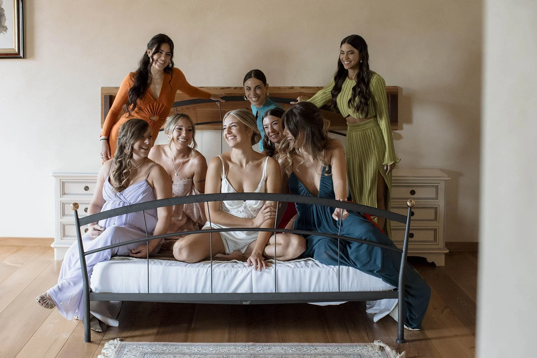 Un gruppo di otto donne vestite con pigiami colorati sedute e in piedi su un letto, che sorridono e si divertono insieme in una stanza.