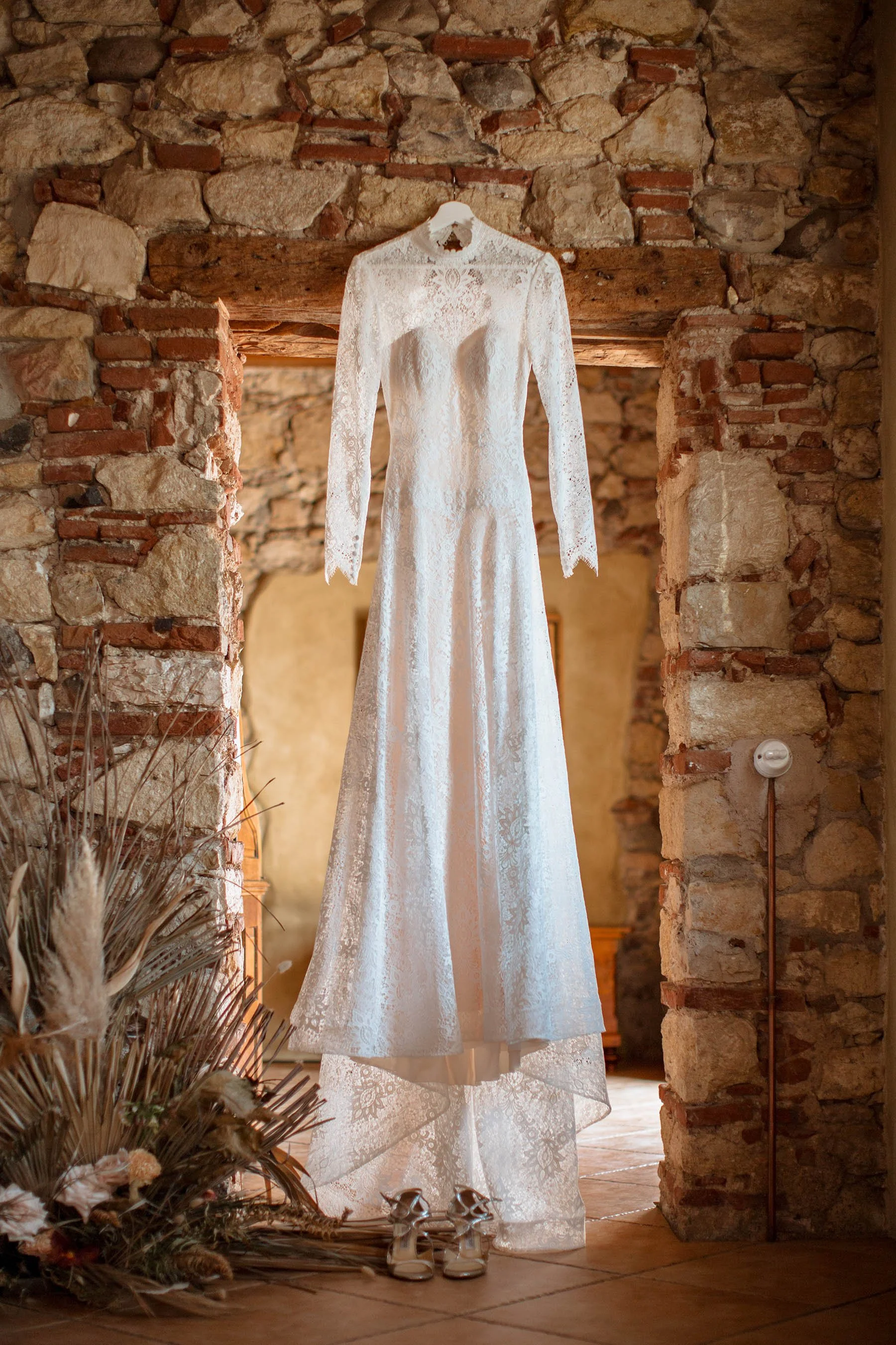 Vestito da sposa bianco in pizzo appeso in una stanza con pareti di pietra e mattoni, con scarpe da sposa in primo piano.