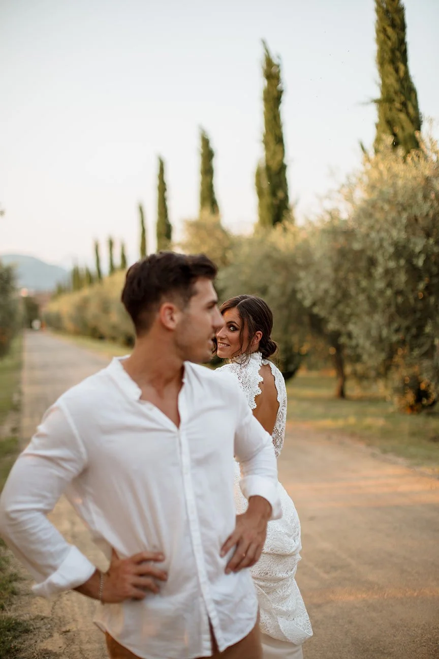 Una coppia si trova su un sentiero nel parco, con alberi e cipressi sullo sfondo, durante il tramonto, con un uomo in camicia bianca e una donna in abito bianco di pizzo.