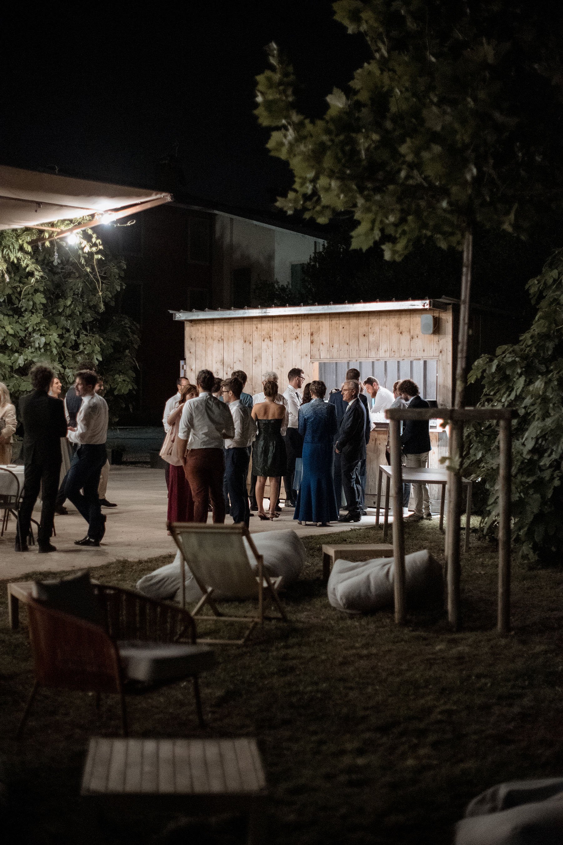 Persone che socializzano e brindano in un evento serale all'aperto, con un'ambientazione rilassata e casual, vicino a una struttura di legno