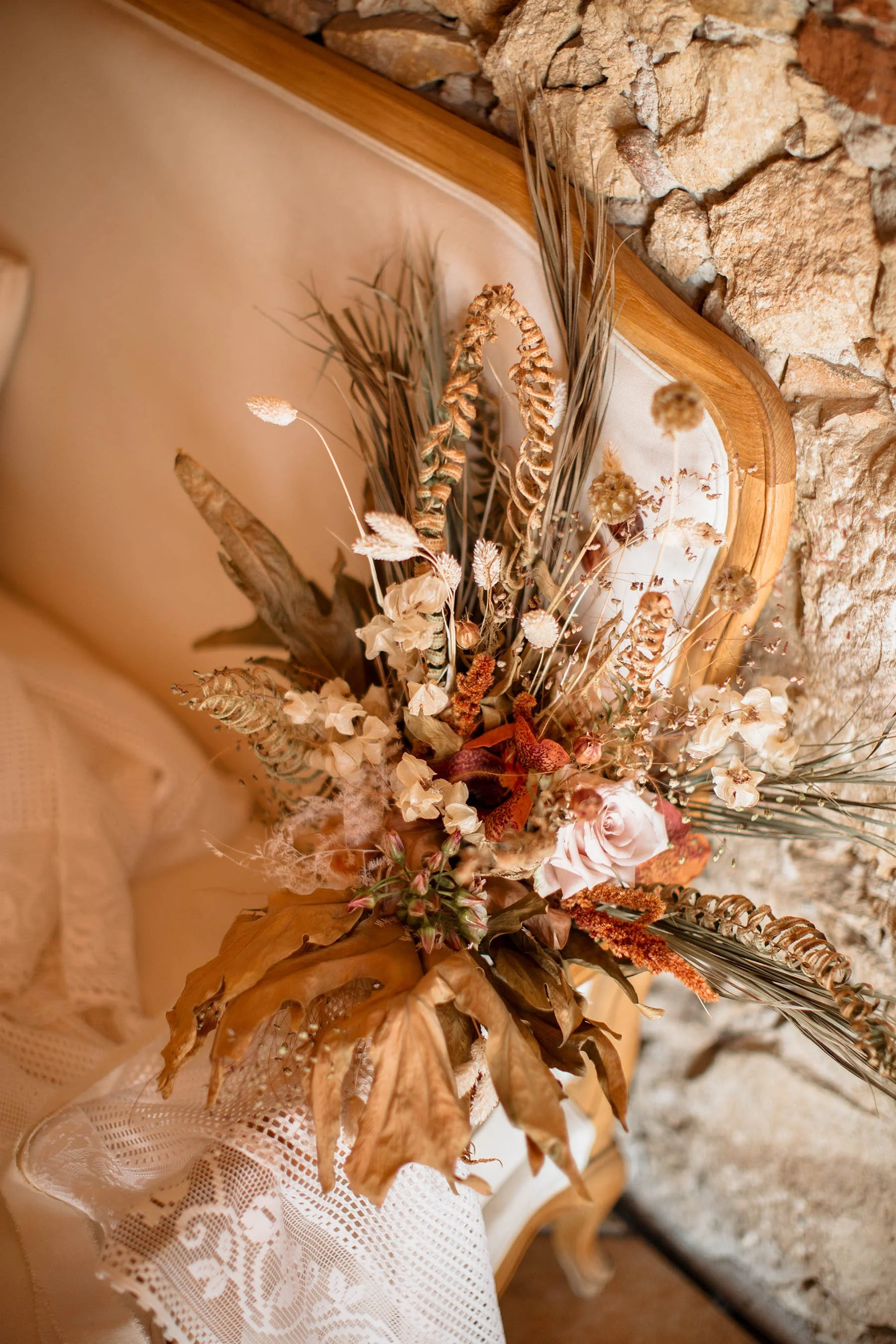 Bouquet di fiori secchi con foglie marroni, graminacee e rose, posato su una sedia in legno con schienale imbottito beige e rivestimento in tessuto, vicino a un muro di pietre.
