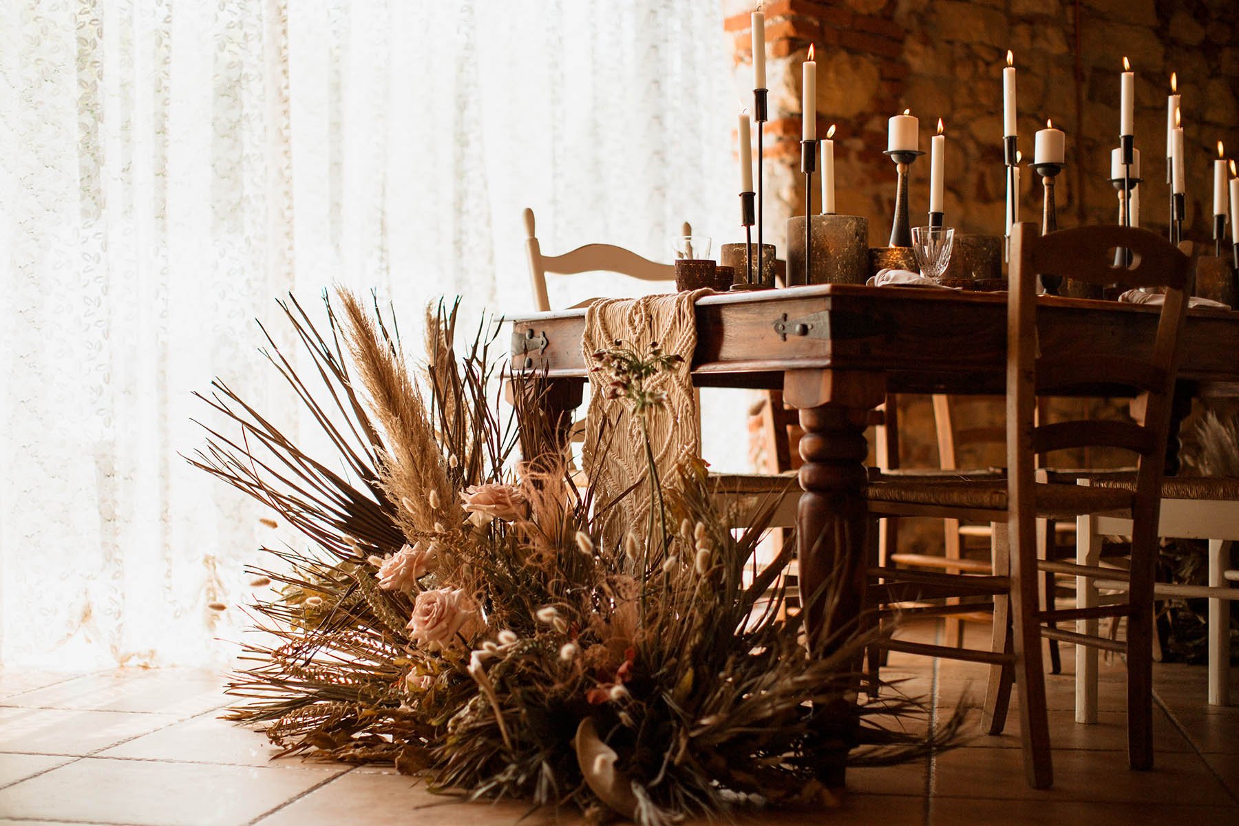 Tavola rustica decorata con candelabri con candele accese, un tovagliolo ricamato, e un grande bouquet di fiori e piante secche posizionato sul pavimento.