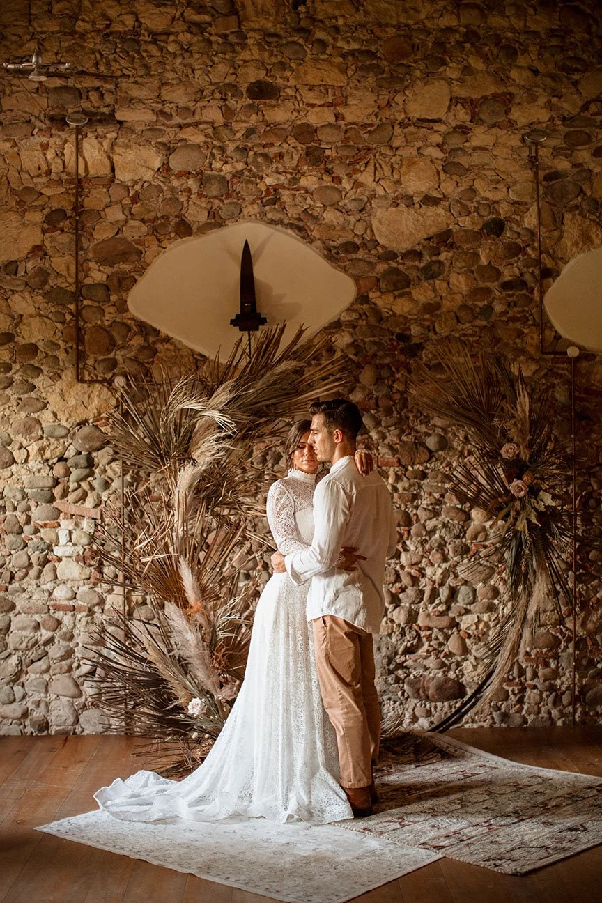 Una coppia si abbraccia in un ambiente rustico con parete in pietra, decorazioni con piante secche e arazzi, in un contesto intimo e romantico.