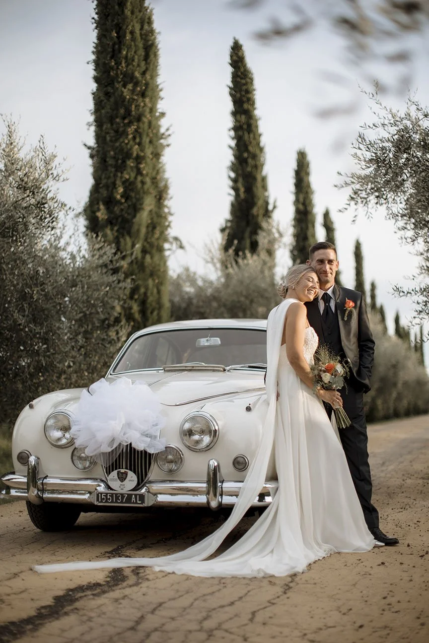 Una coppia di sposi davanti a un'auto d'epoca decorata con nastro bianco, con uno sfondo di alberi di cipresso e altri arbusti, durante un matrimonio.