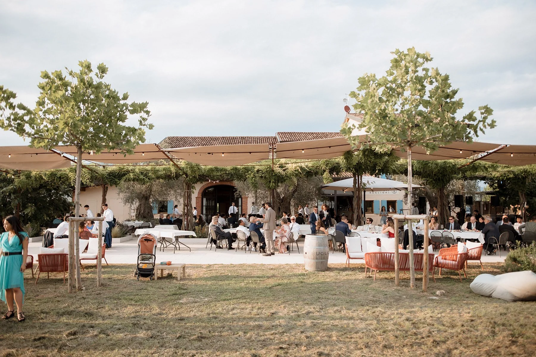 Evento di cerimonia all'aperto con persone sedute a tavoli, alcuni in abbigliamento formale, sotto tende e con alberi decorativi.