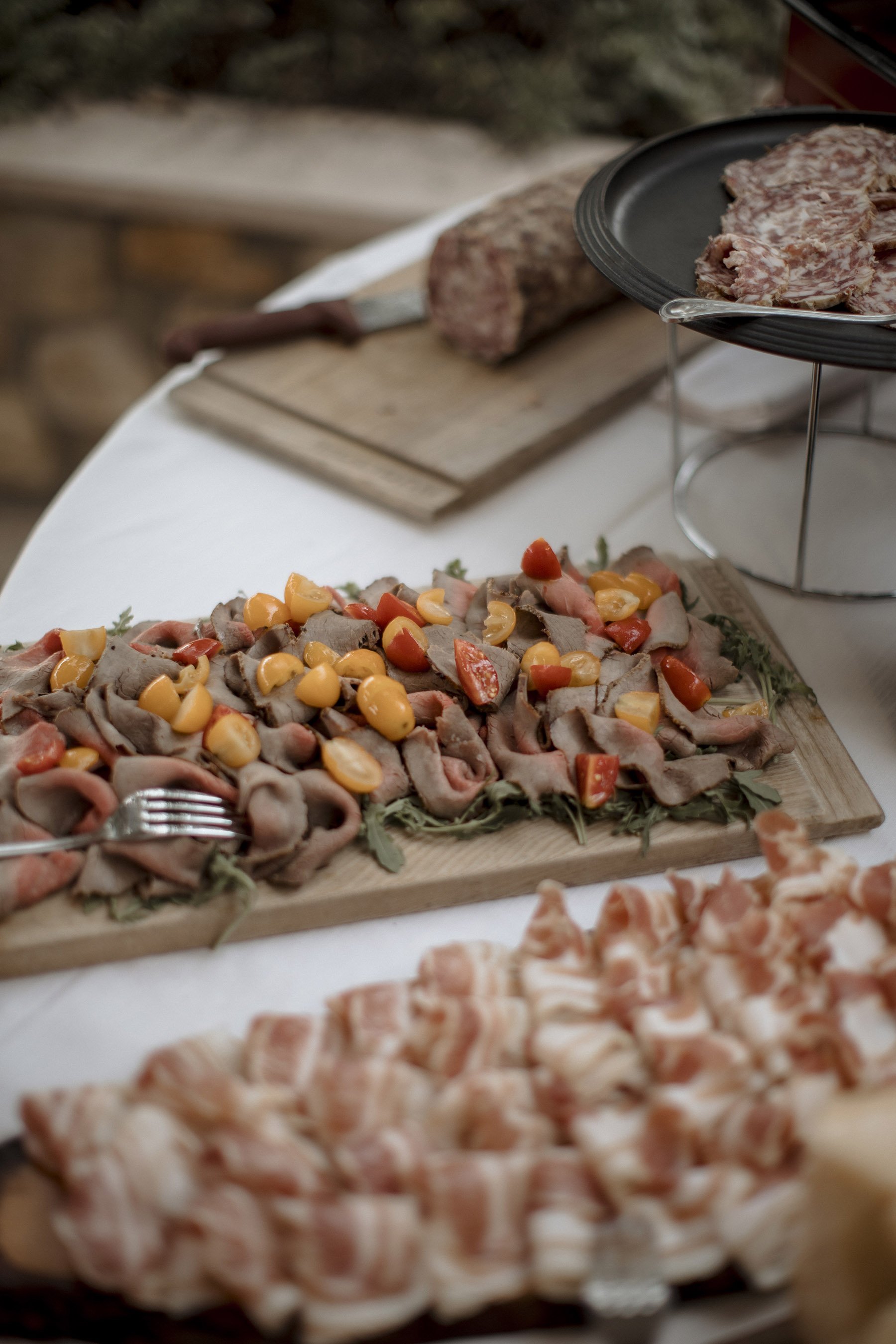 Assorted cold cuts and cured meats arranged on a wooden platter, with cherry tomatoes and greens, on a white tablecloth.