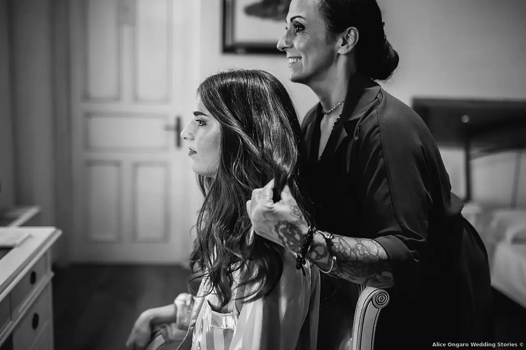 Una donna anziana che si prende cura di una ragazza con lunghi capelli scuri, seduta in una stanza con pareti chiare, in un momento che sembra di cura e attenzione, in bianco e nero.