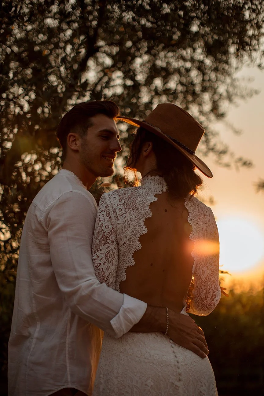 Una coppia felice durante un tramonto, l'uomo tiene la donna per la vita, entrambi sorridenti, con uno sfondo di alberi e il sole al tramonto.