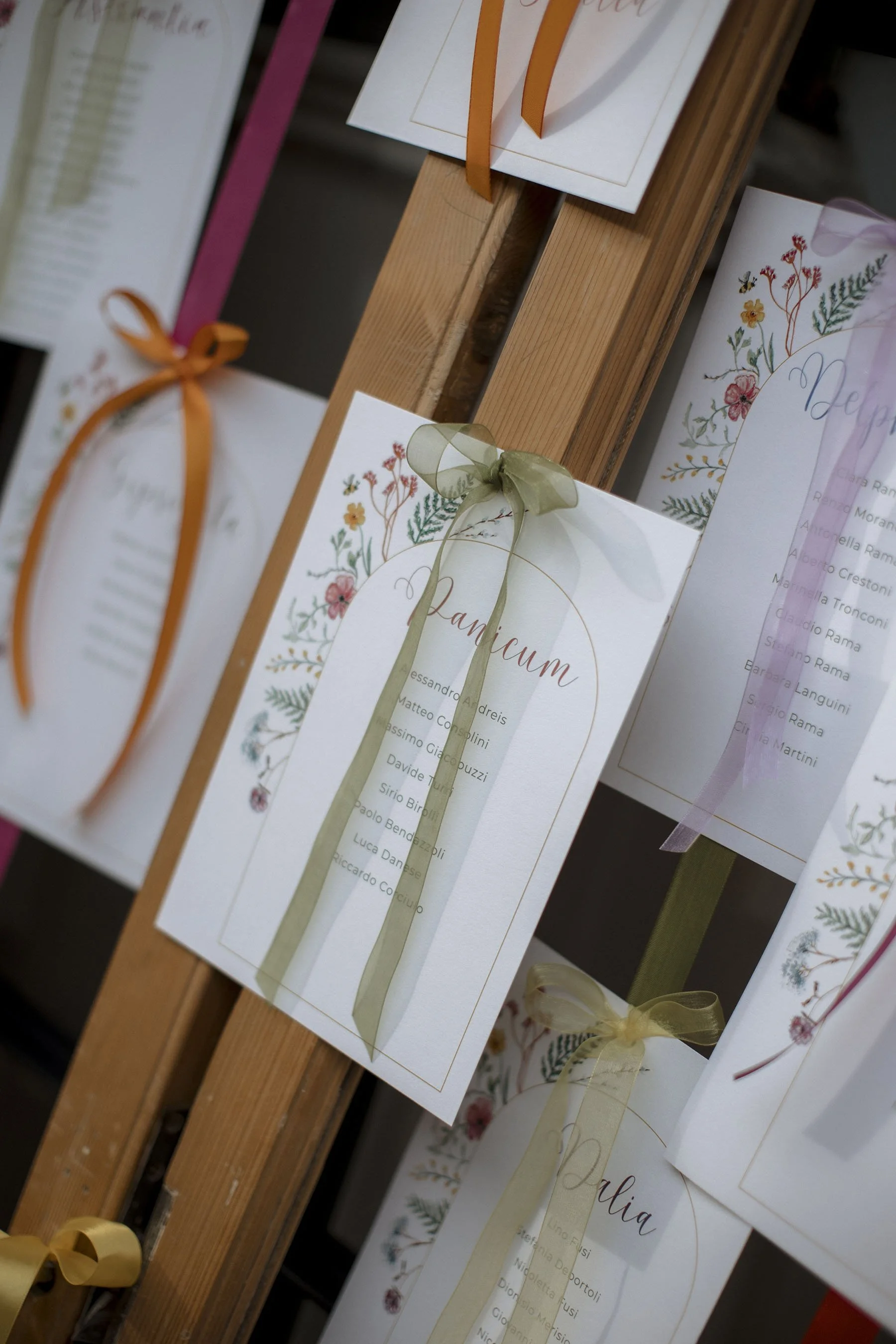 Cartellini floreali e rigati con liste di nomi, appesi con fiocchi di raso, che sembrano essere menù o programmi per un evento speciale, come un matrimonio o una festa.