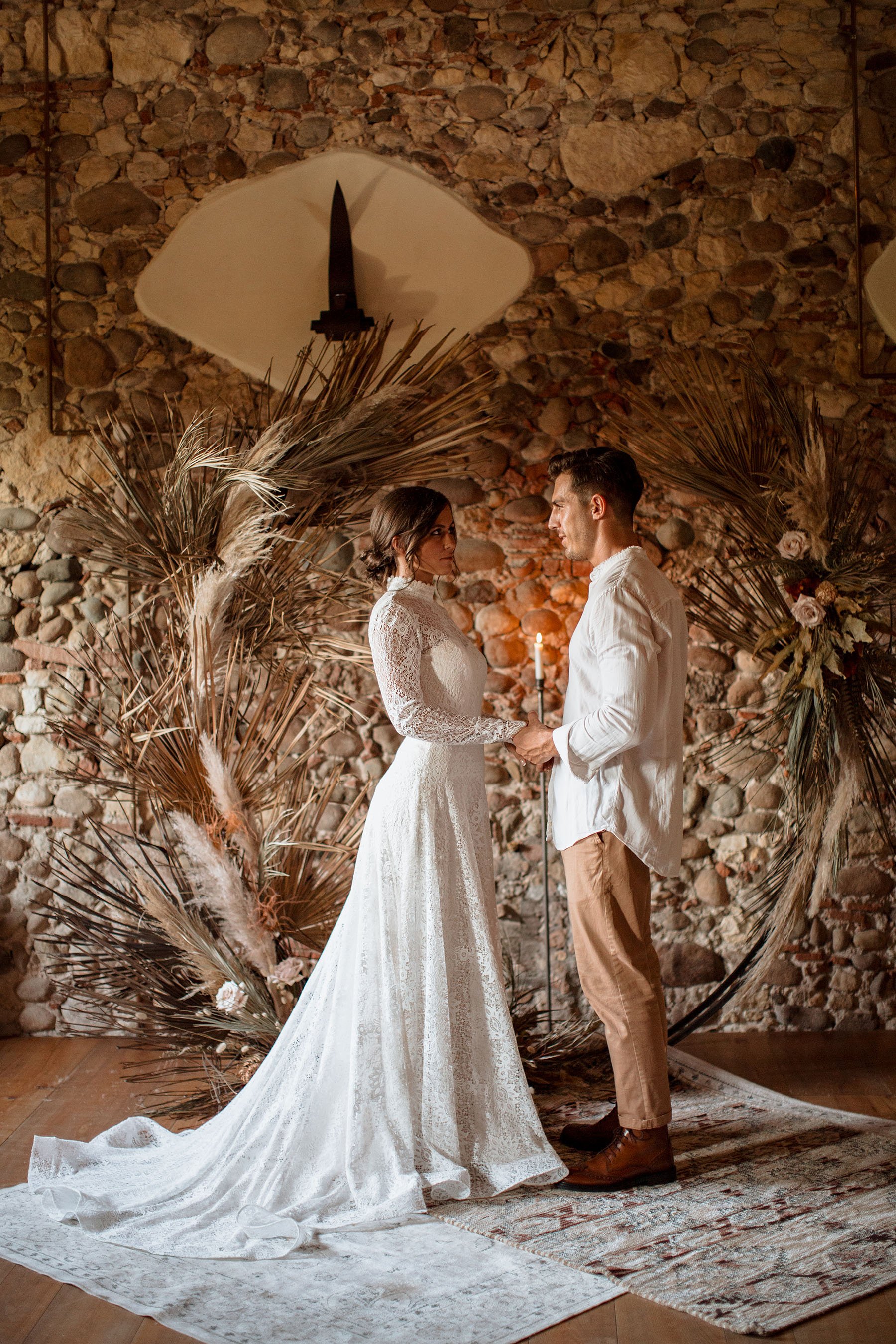 Una coppia si tiene per mano durante una cerimonia di matrimonio in un ambiente rustico con mura di pietra. La sposa indossa un abito bianco in pizzo con lunghe maniche, mentre lo sposo indossa una camicia bianca e pantaloni beige. Sullo sfondo, deco