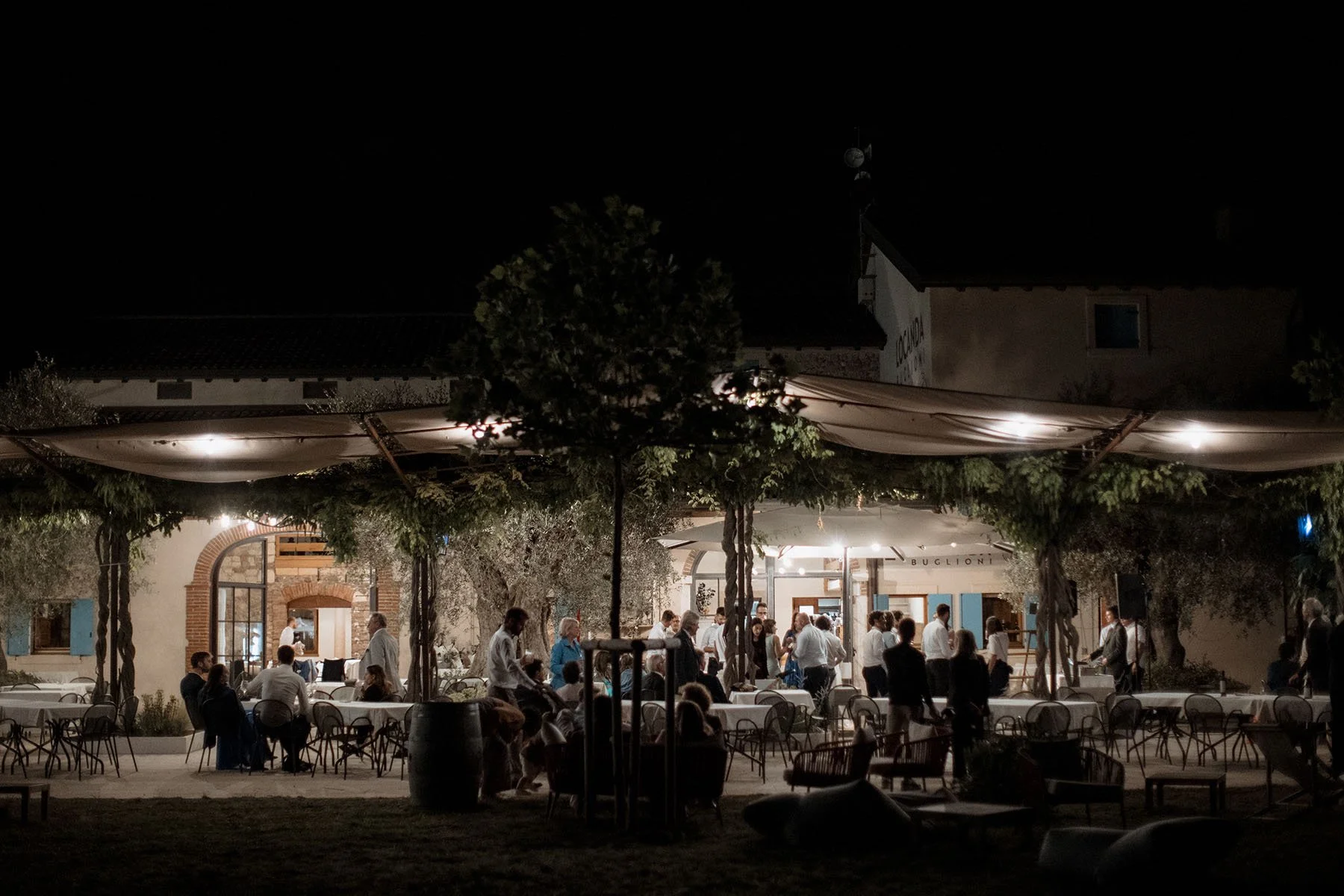 Festa all'aperto con persone che socializzano sotto luci soffuse, tavoli e ombrelloni in un ambiente serale, con alberi e un edificio sullo sfondo.