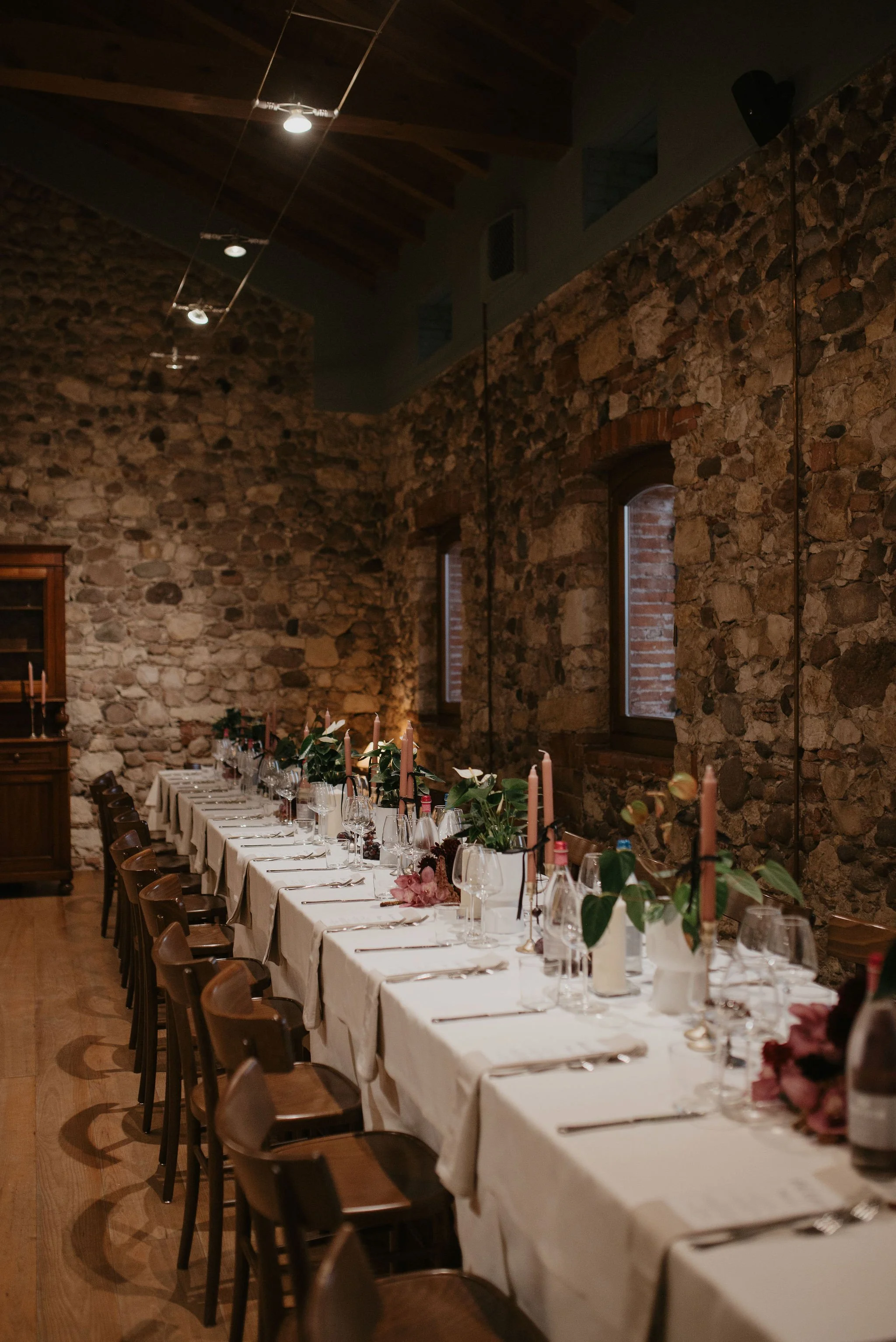 Tavola apparecchiata per un banchetto con tovaglia bianca, candelabri con candele rosa, vasi con fiori e bicchieri di vetro su un ambiente rustico con pareti di pietra e finestre.