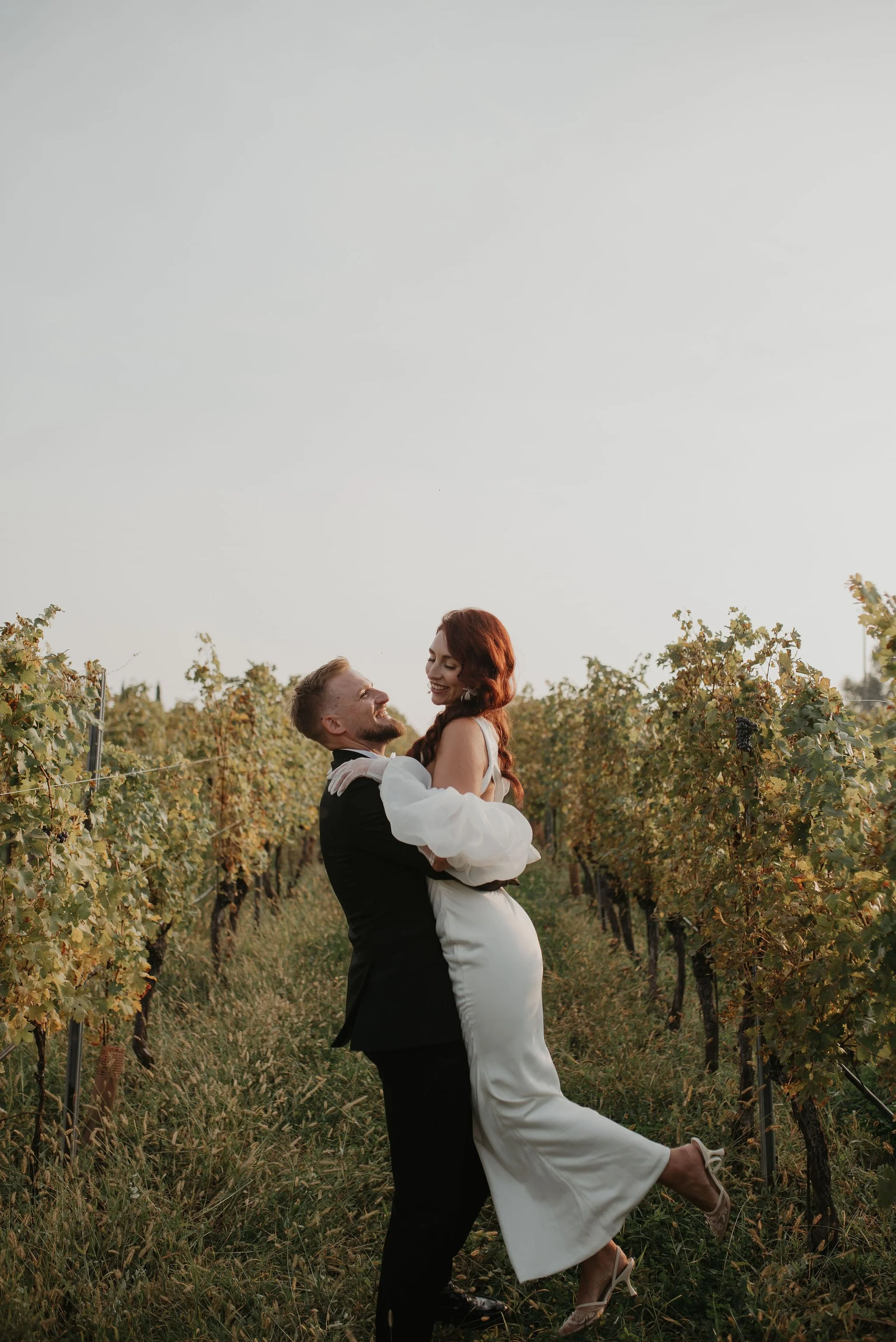 Una coppia felice in abiti eleganti, di fronte a un vigneto, si abbraccia e ride, al tramonto.