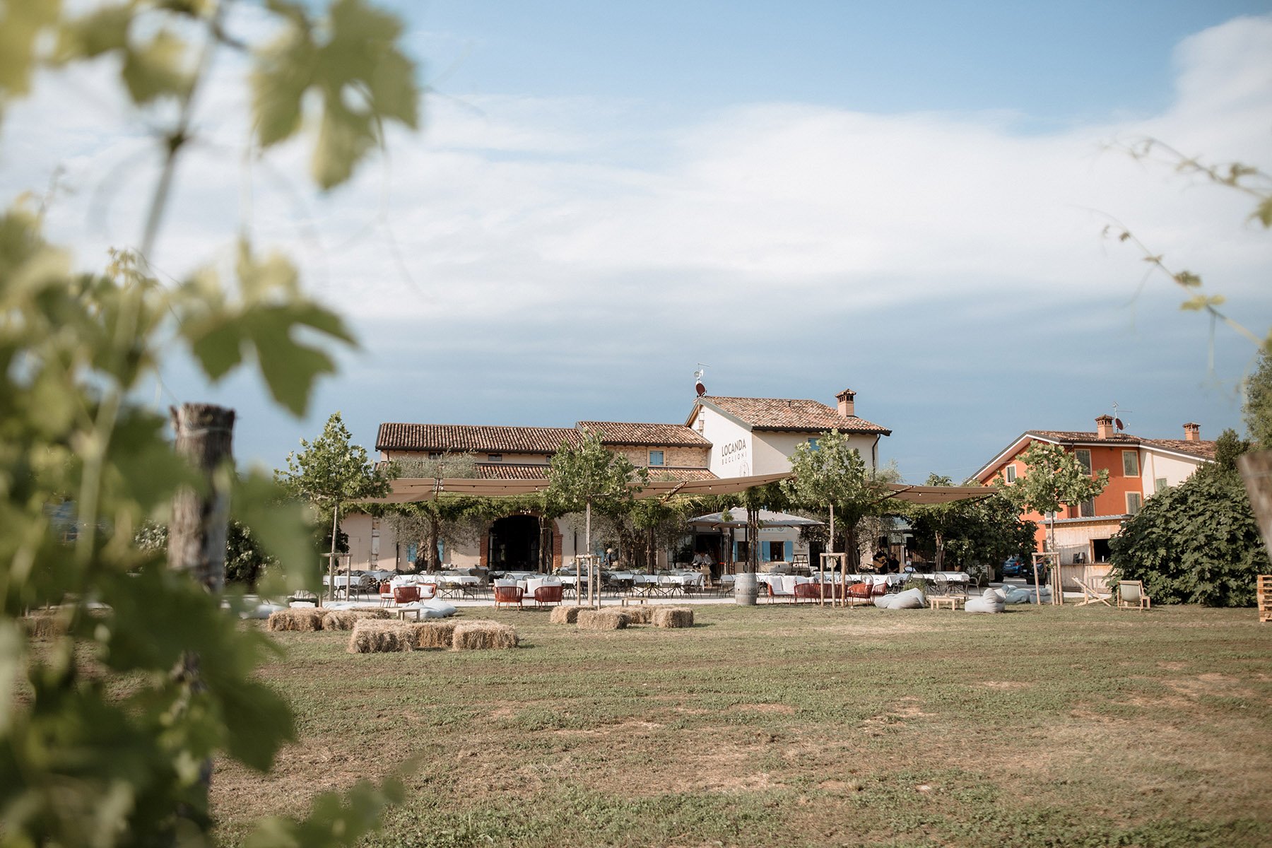 Area all'aperto con tavoli, sedie e balle di fieno davanti a un ristorante, circondato da qualche albero e case in stile rurale.