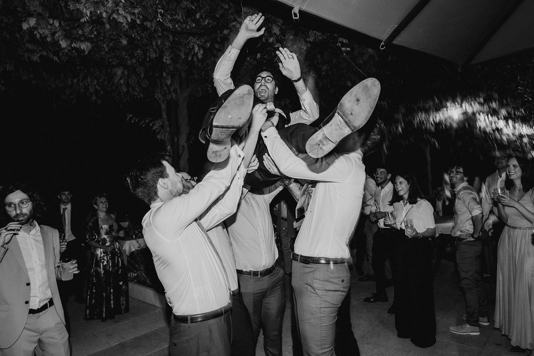 Un gruppo di uomini sollevano un uomo seduto mentre ride durante una festa all'aperto di notte.