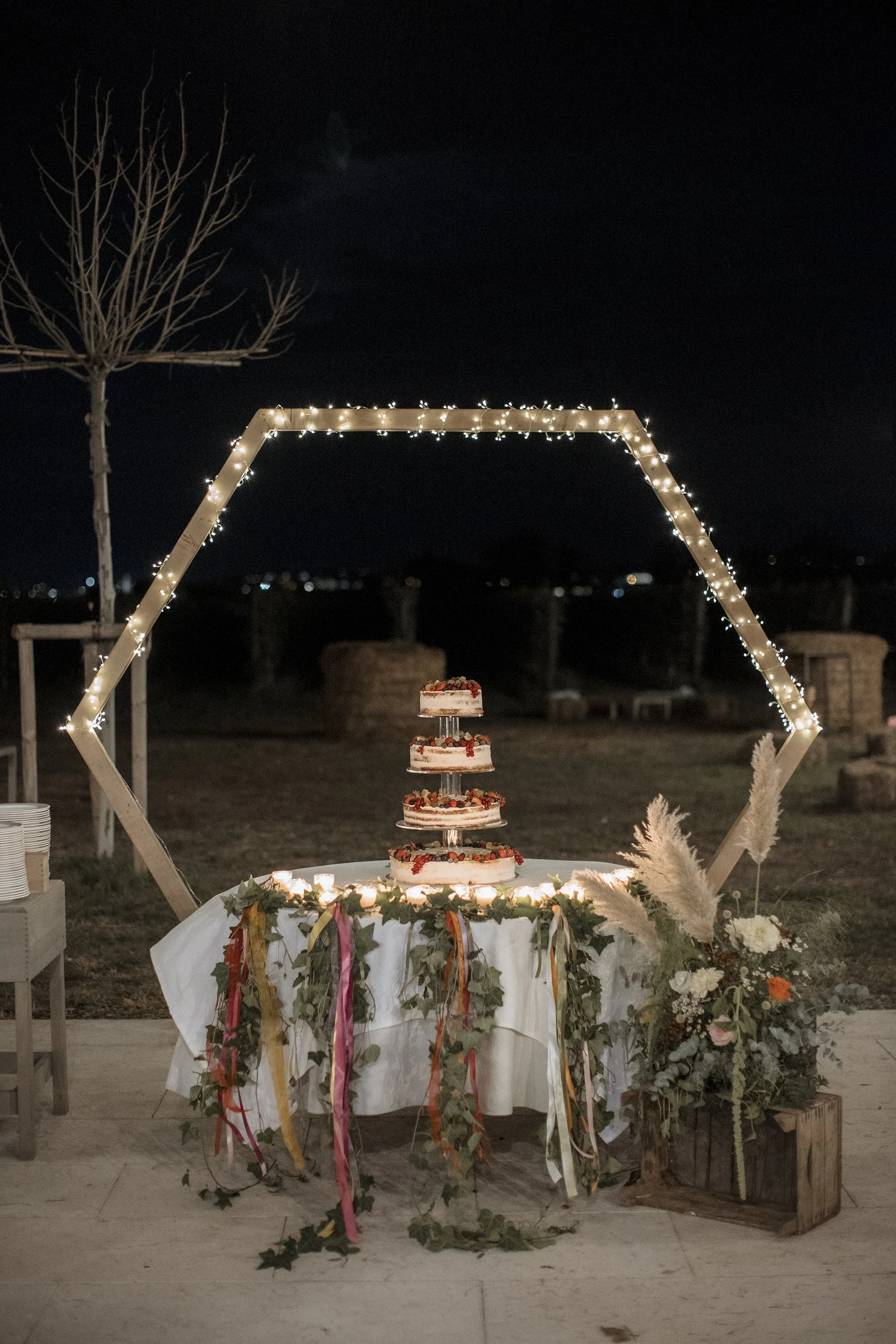 Tavolo decorato con torta a più piani, illuminato da luci natalizie. Dietro, un'installazione di legno a forma di esagono e un albero senza foglie. Scenografia di sera con sfondo scuro e alcune balle di paglia o pietre.