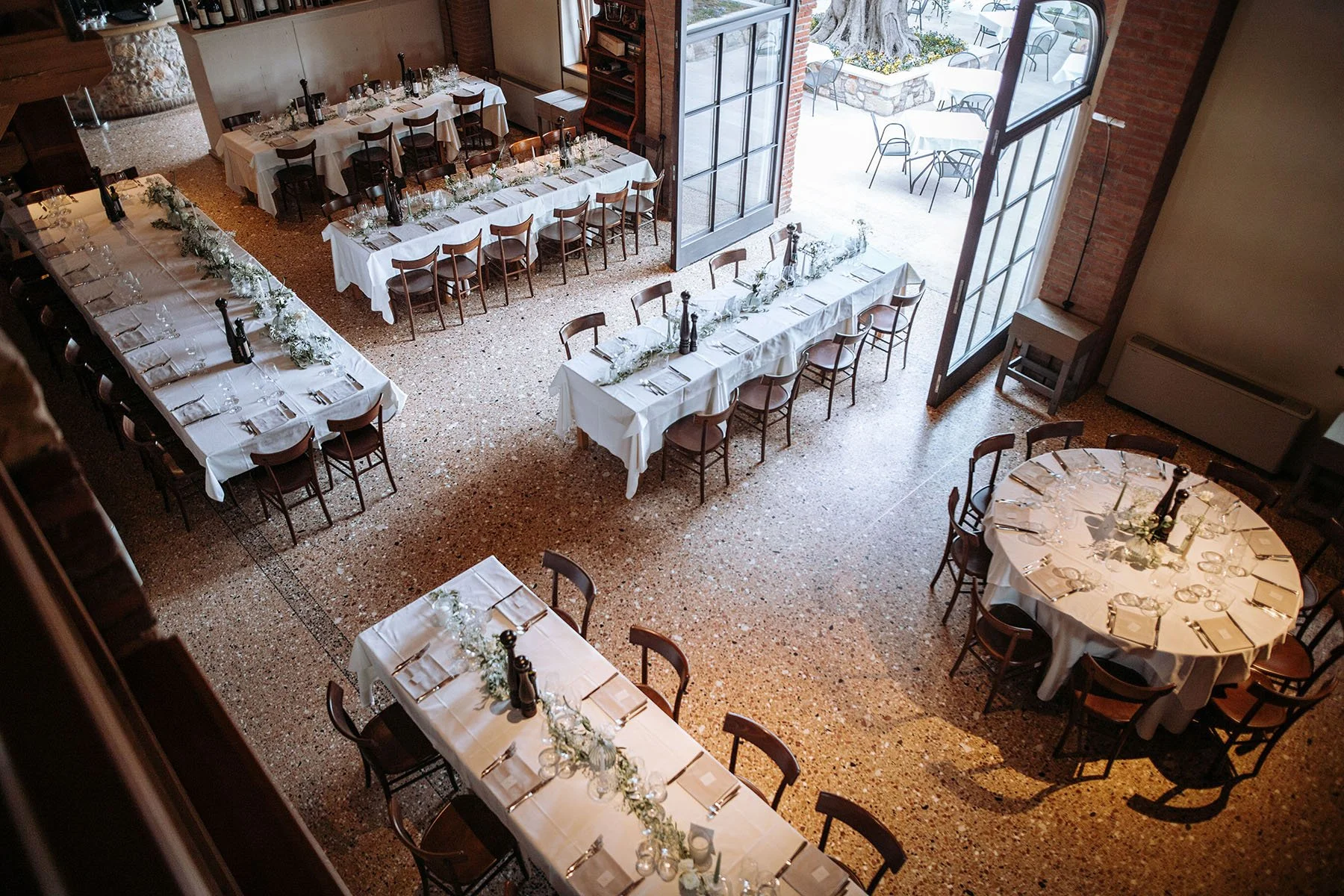 Sala per un evento o ricevimento con tavoli apparecchiati con tovaglie bianche, decorazioni floreali e bottiglie di vino. Grande porta aperta che dà su un patio esterno con tavolini e sedie.