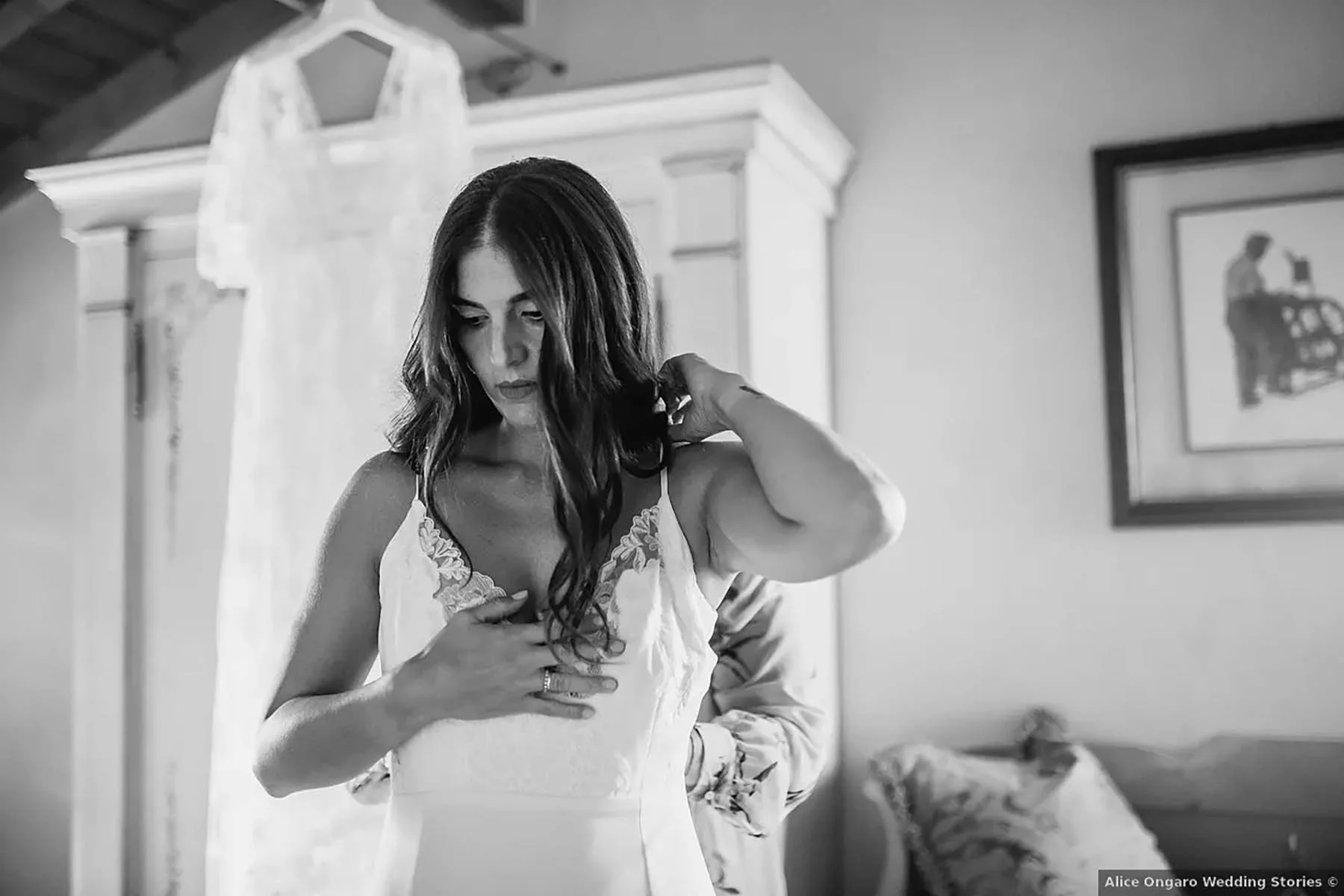 Una donna con capelli castani scuri si sta aggiustando il vestito, che sembra essere un abito da sposa, in una stanza con mobili classici e un quadro sul muro.