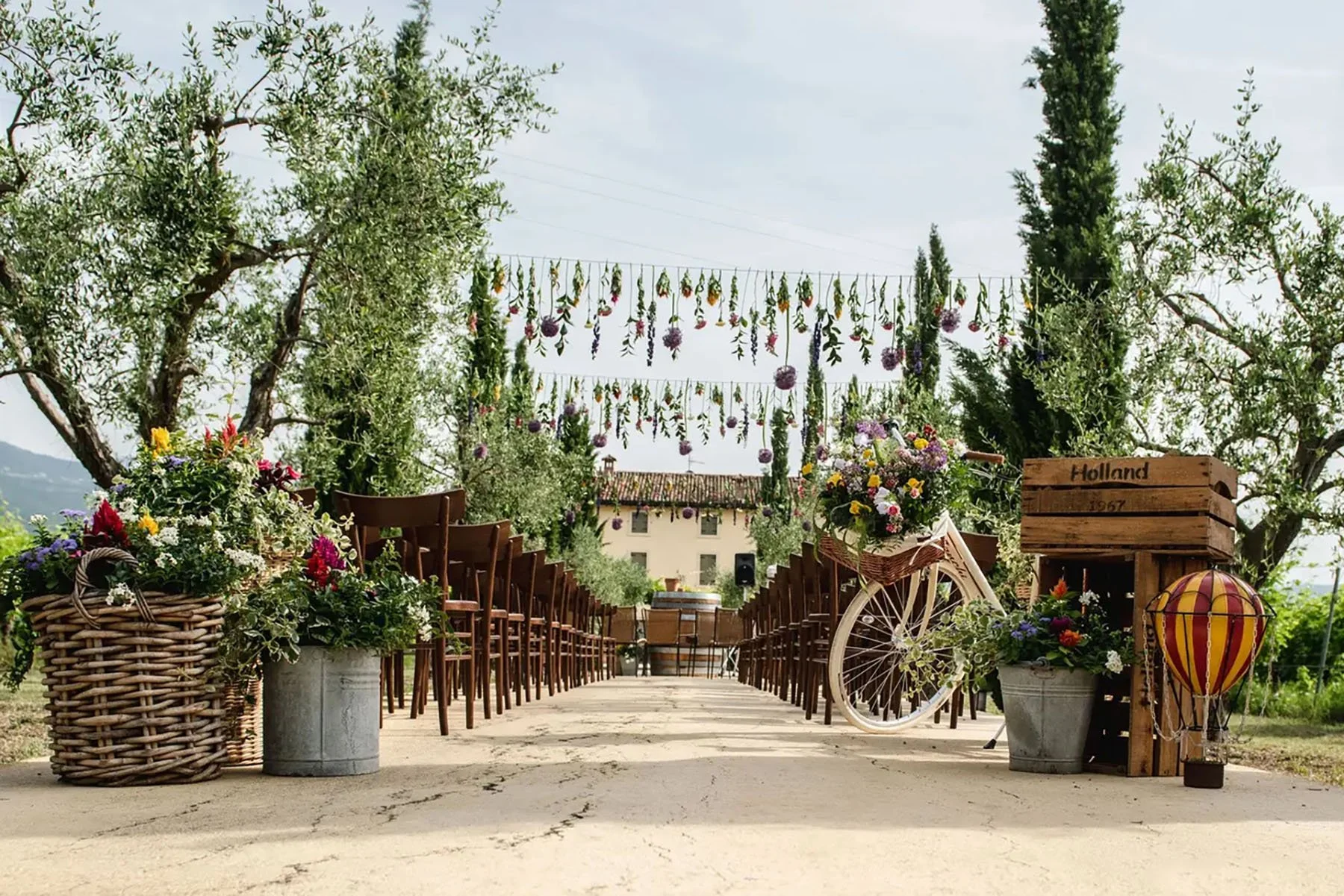 Decorazione per un matrimonio su una strada, con sedie di legno ai lati, fiori, cestini, un carro a bici con fiori,ed è una scena all'aperto tra alberi e un edificio di fondo.