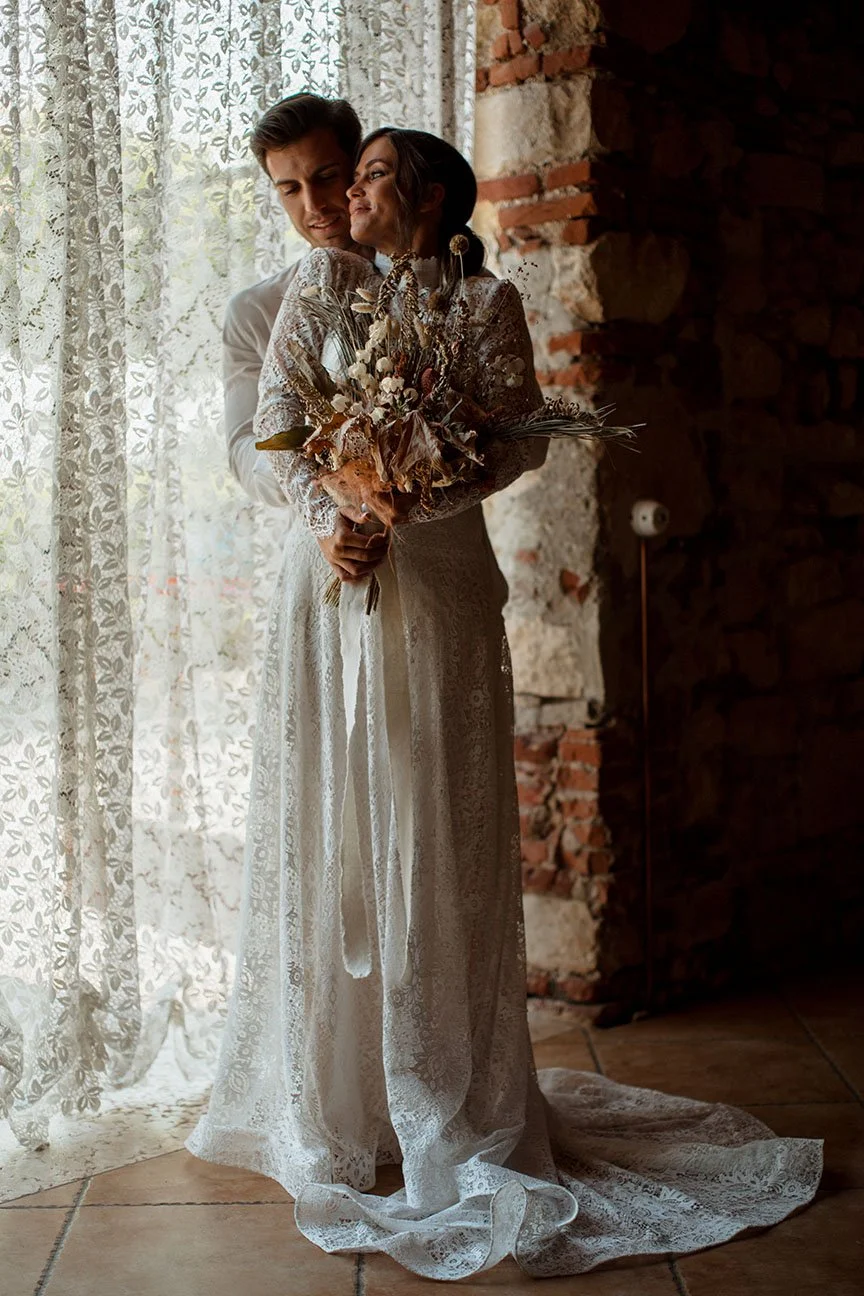 Una coppia di sposi in abiti da matrimonio, con la donna che tiene un bouquet di fiori secchi, si abbracciano dolcemente davanti a una finestra con tende di pizzo, in un ambiente rustico con pareti di pietra e mattoni.