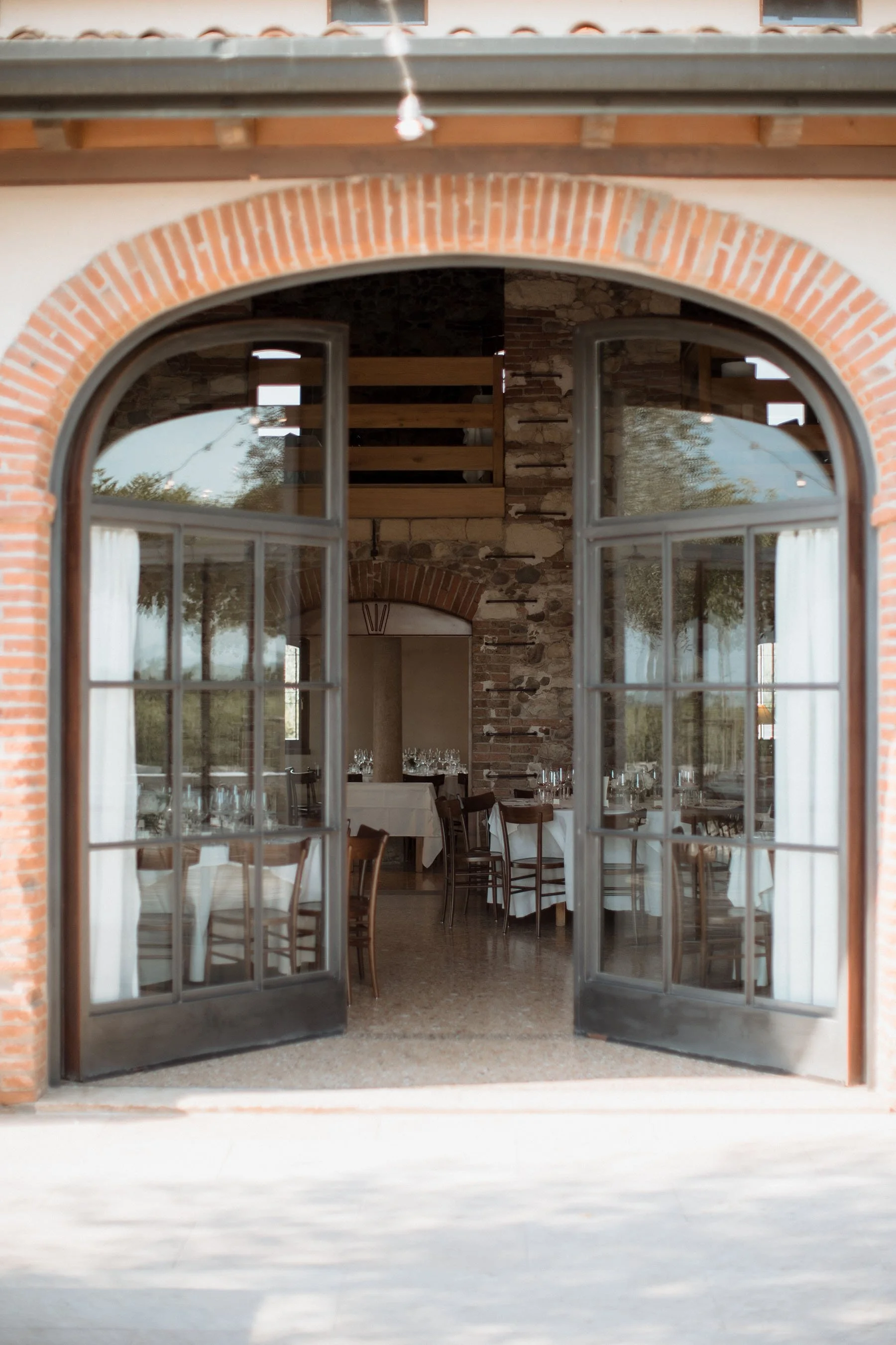 Interno di un ristorante visto attraverso una porta a vetri con archi in mattoni, con tavoli apparecchiati con tovaglie bianche e bicchieri, pareti in pietra e decorazioni rustiche.