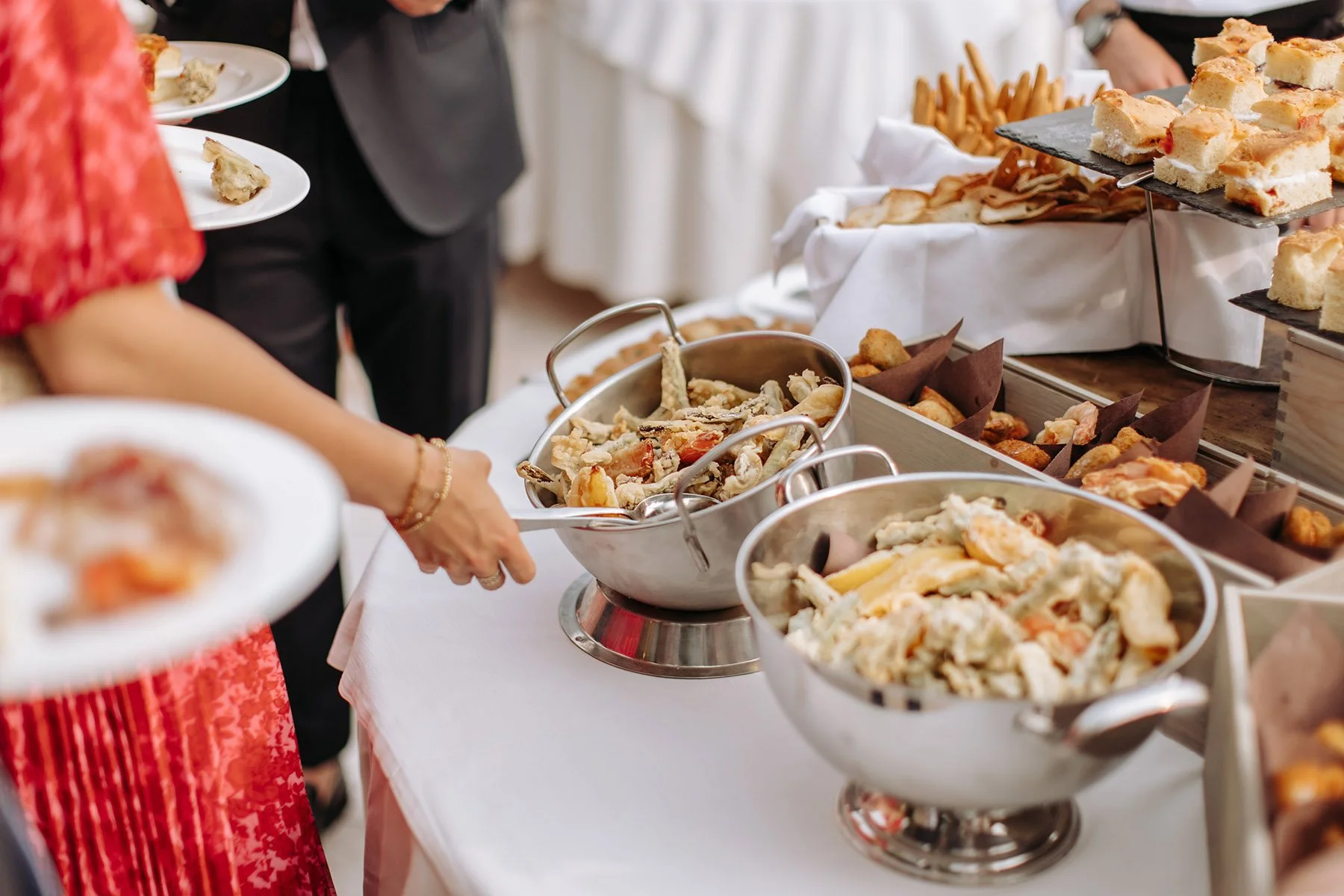 Buffet con vari tipi di alimenti, tra cui insalate, fritti, pane e affettati, con persone che si servono.