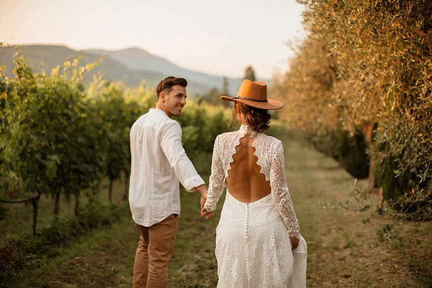 Una coppia di persone felici si tiene per mano in un vigneto durante il tramonto, indossando abiti estivi eleganti.