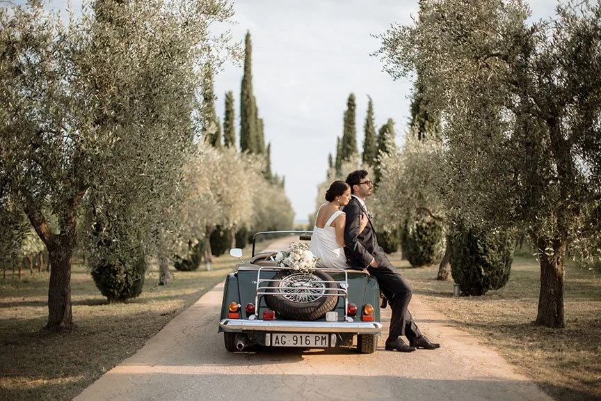 Una coppia di sposi seduta sul retro di un'auto decorata con fiori, che attraversa un viale di alberi, forse in un contesto di matrimonio o romantico.