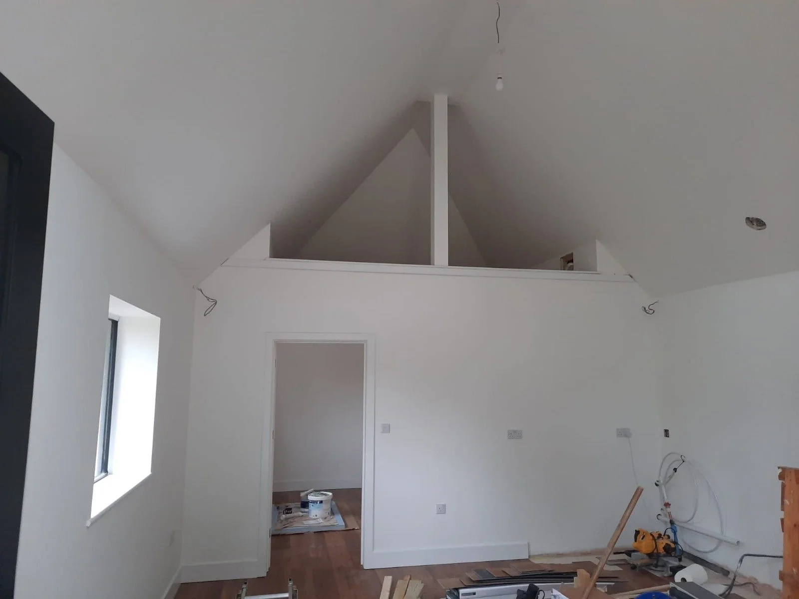 Interior of a house under construction, with white walls and an angled ceiling, visible wiring, and construction tools and materials on the floor.