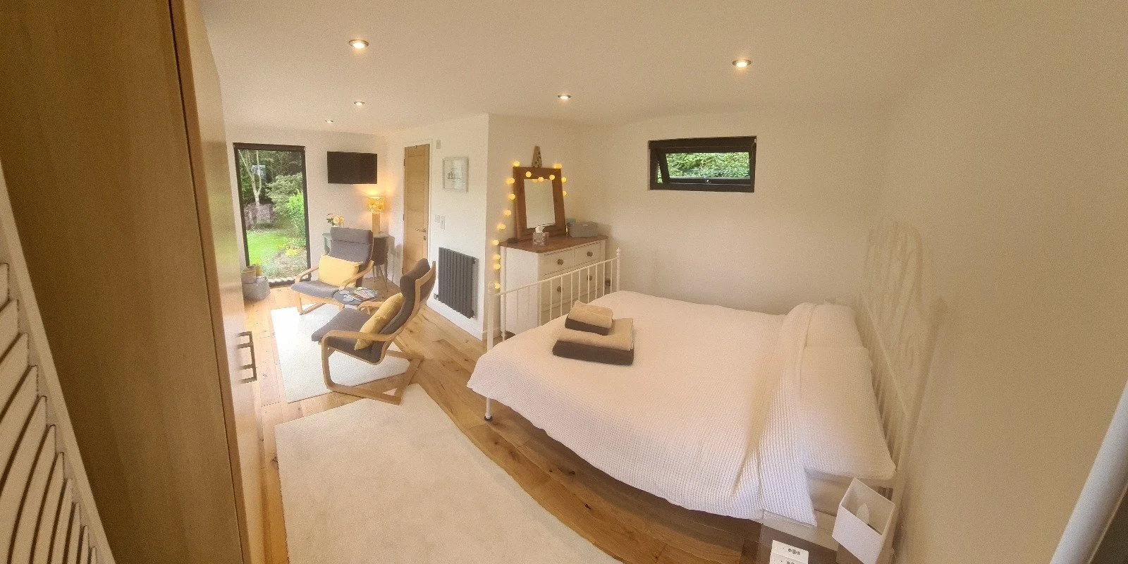 A cozy, modern bedroom with a white bed, stacked towels, wooden flooring, and minimalistic decor. There's a sitting area with chairs and a small table, a window, and soft lighting creating a warm atmosphere.