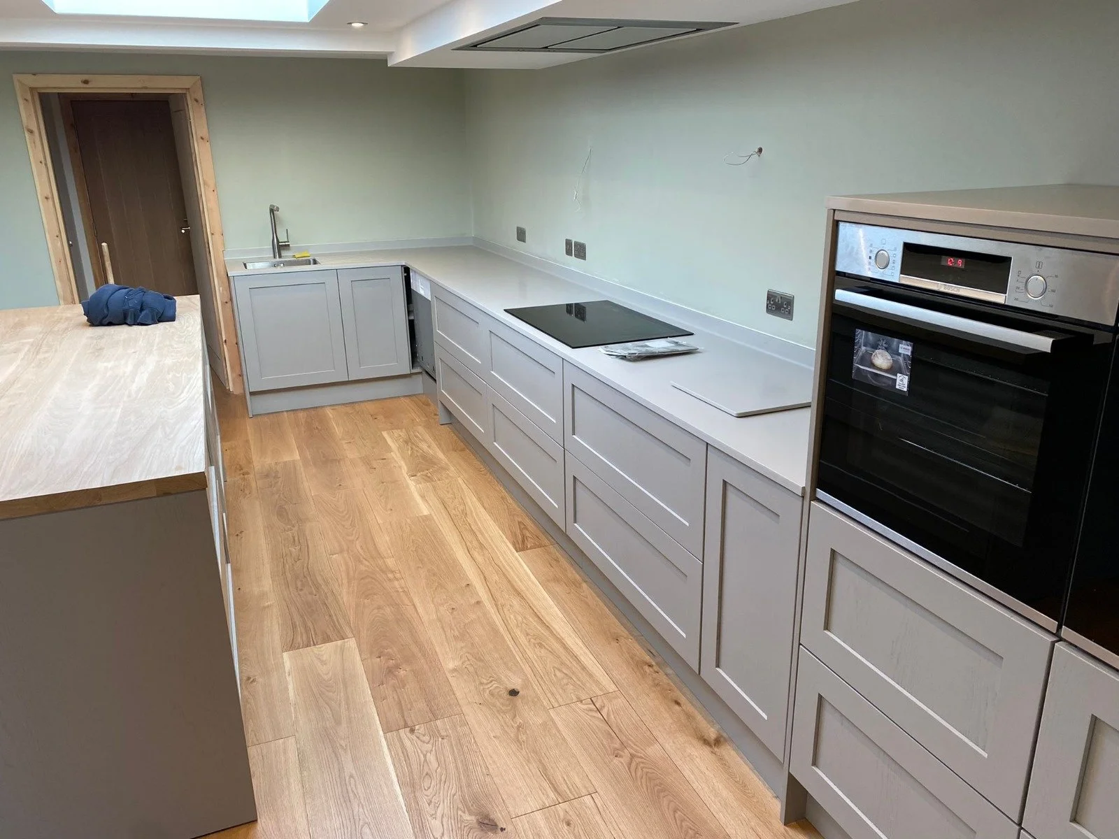Kitchen under construction with grey cabinets, wooden flooring, a built-in oven, an electric stovetop, and a sink, with some tools and materials on the countertops.