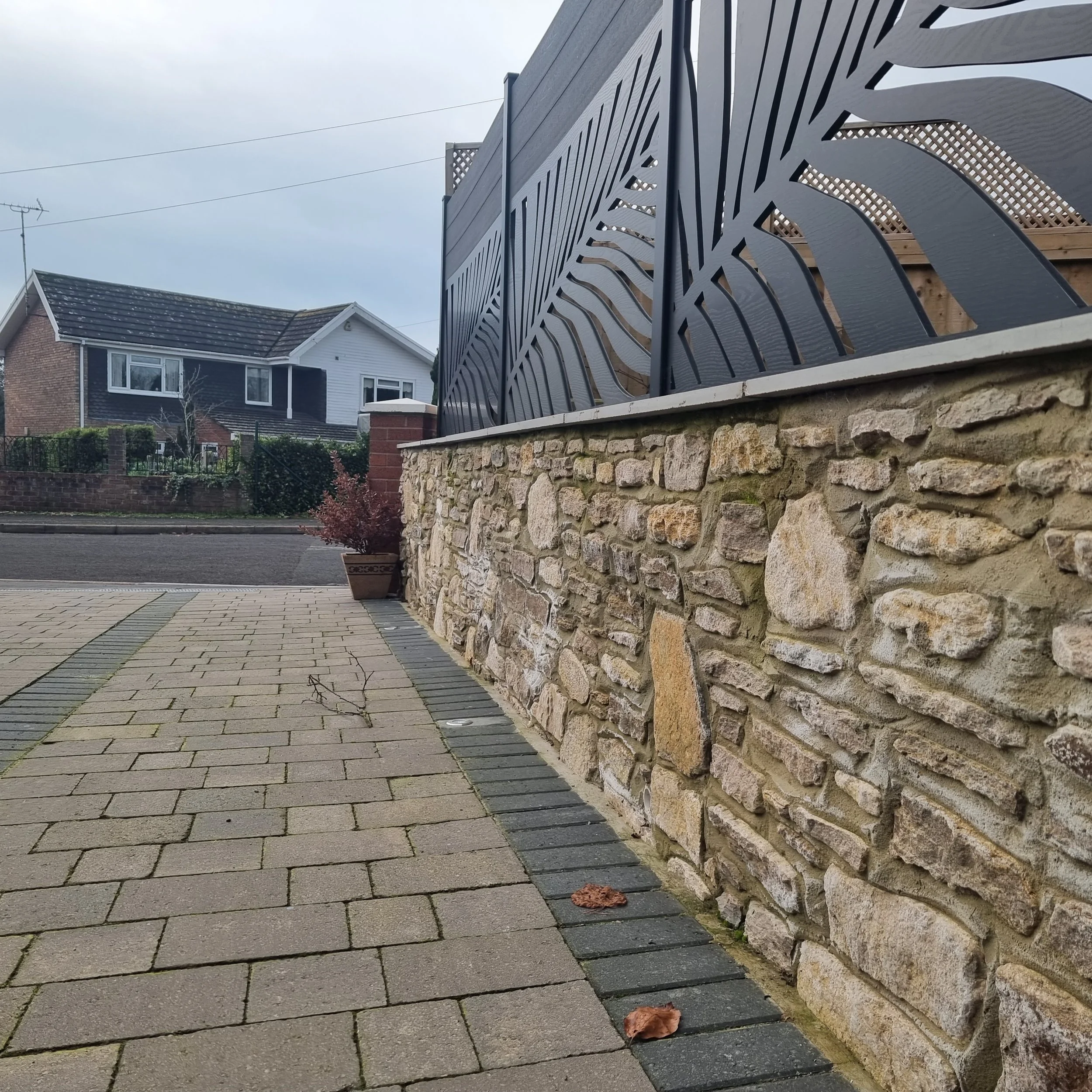 Sidewalk and stone wall with decorative metal fencing on top, with a residential house and plants in the background.