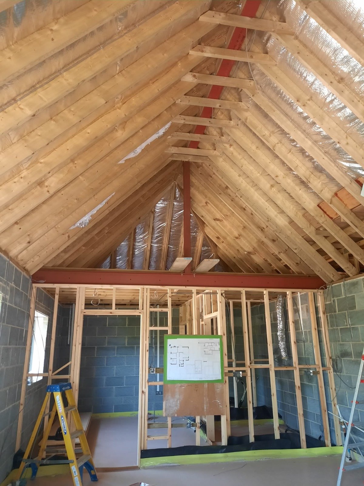 Interior view of a house under construction showing an unfinished roof with wooden framing and insulation, and a partially built wall structure with a floor plan diagram on a board.