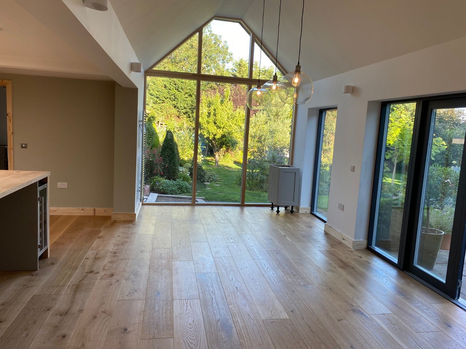 A spacious room with large floor-to-ceiling windows and sliding glass doors, wooden flooring, and a view of a lush green garden outside, with hanging light bulbs.