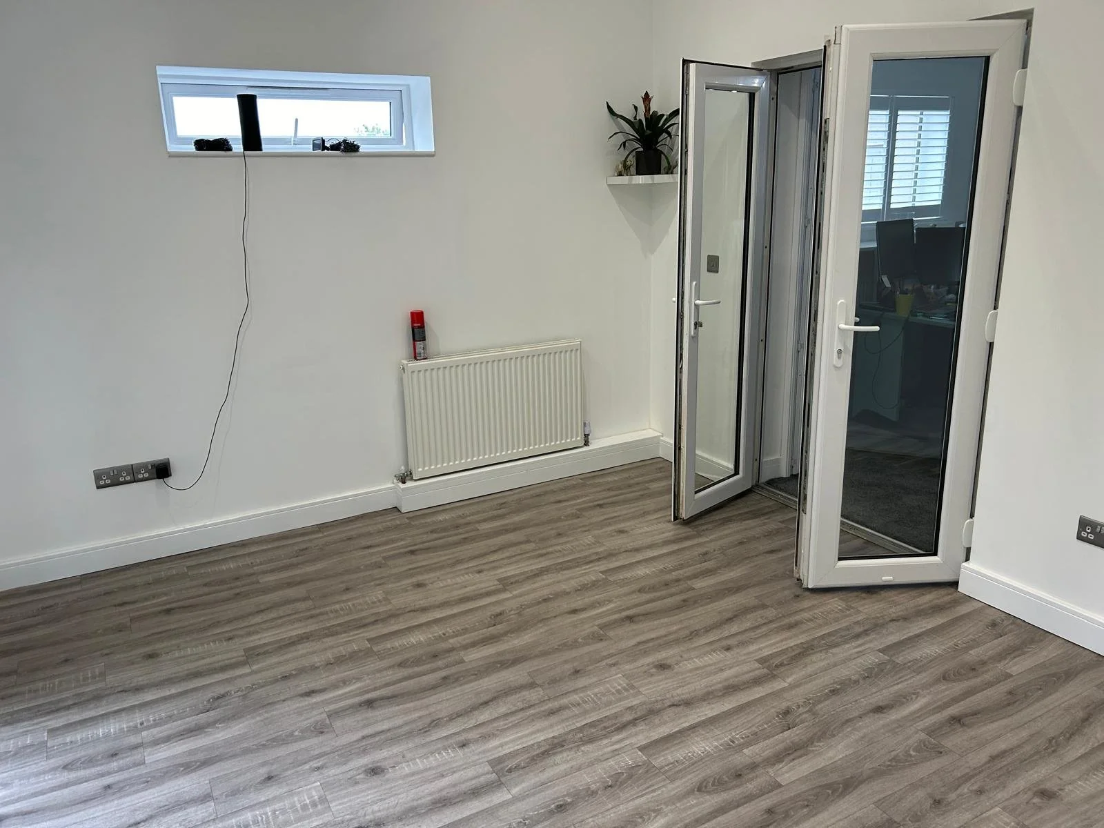 Empty room with wooden flooring, small high window, wall-mounted radiator, power outlets, and glass door with frame.
