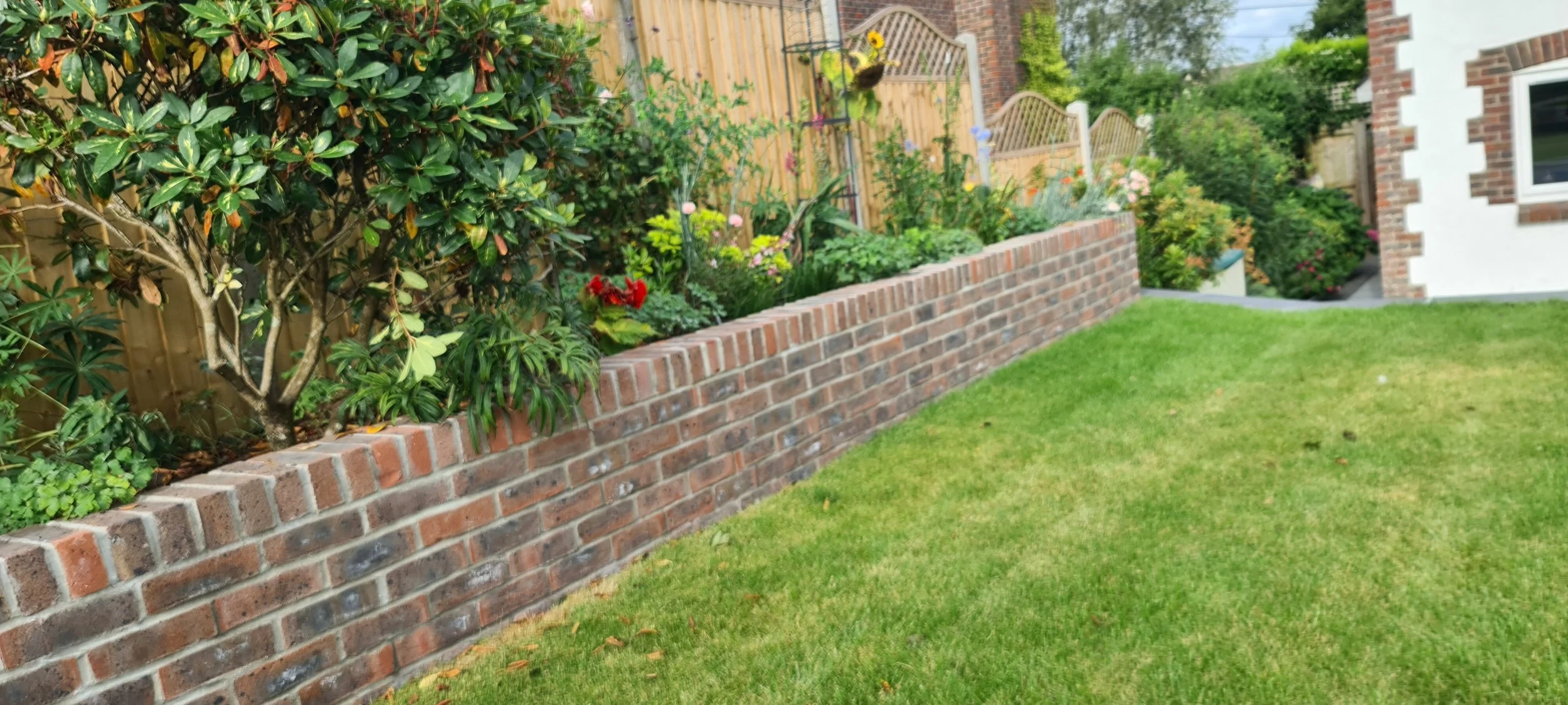 A brick garden bed filled with various plants and flowers, lined with a wooden fence behind it, with a green lawn in the foreground and a white house with brick accents in the background.