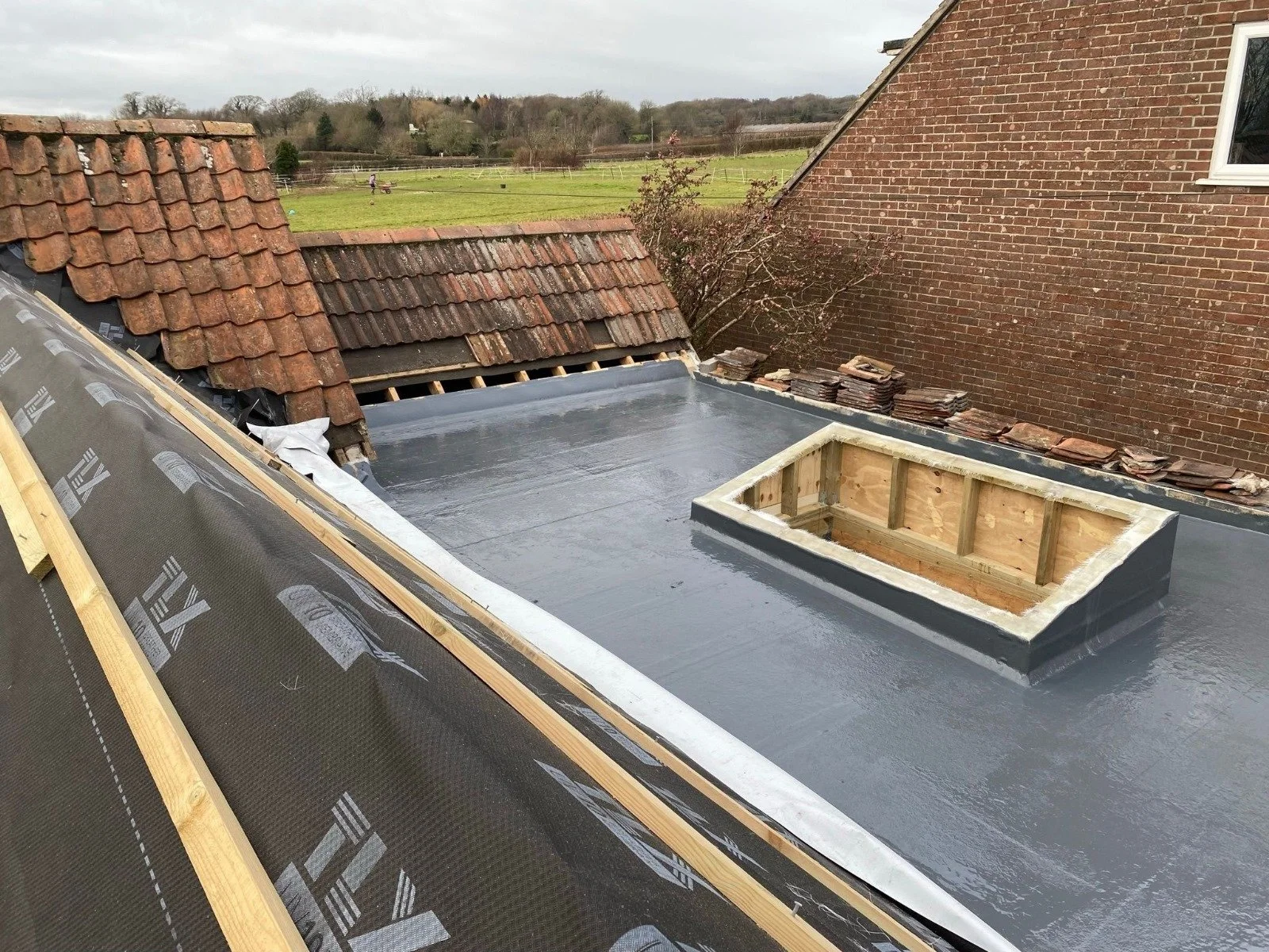 Roof with new waterproofing material and a box-shaped vent opening under construction, with older roof tiles nearby.