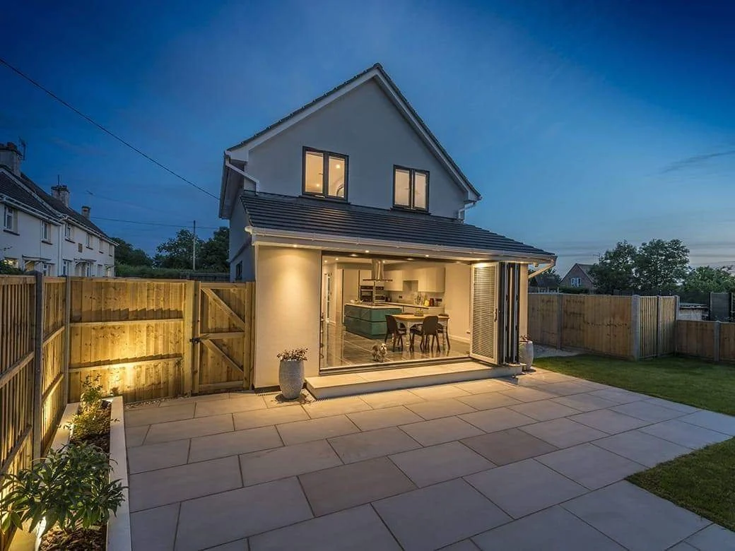 View of a modern two-story house with sliding glass doors open to a backyard patio, illuminated evening scene with backyard fencing, potted plants, and small dog near dining table inside.