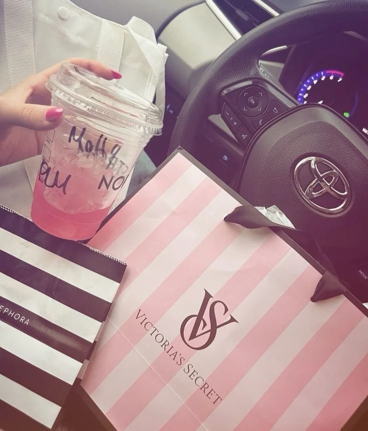 Einkaufstaschen von Victoria's Secret und Sephora, eine Hand hält ein pinkes Getränk in einem transparenten Becher, im Hintergrund ein Auto mit Steuerrad und Armaturen.