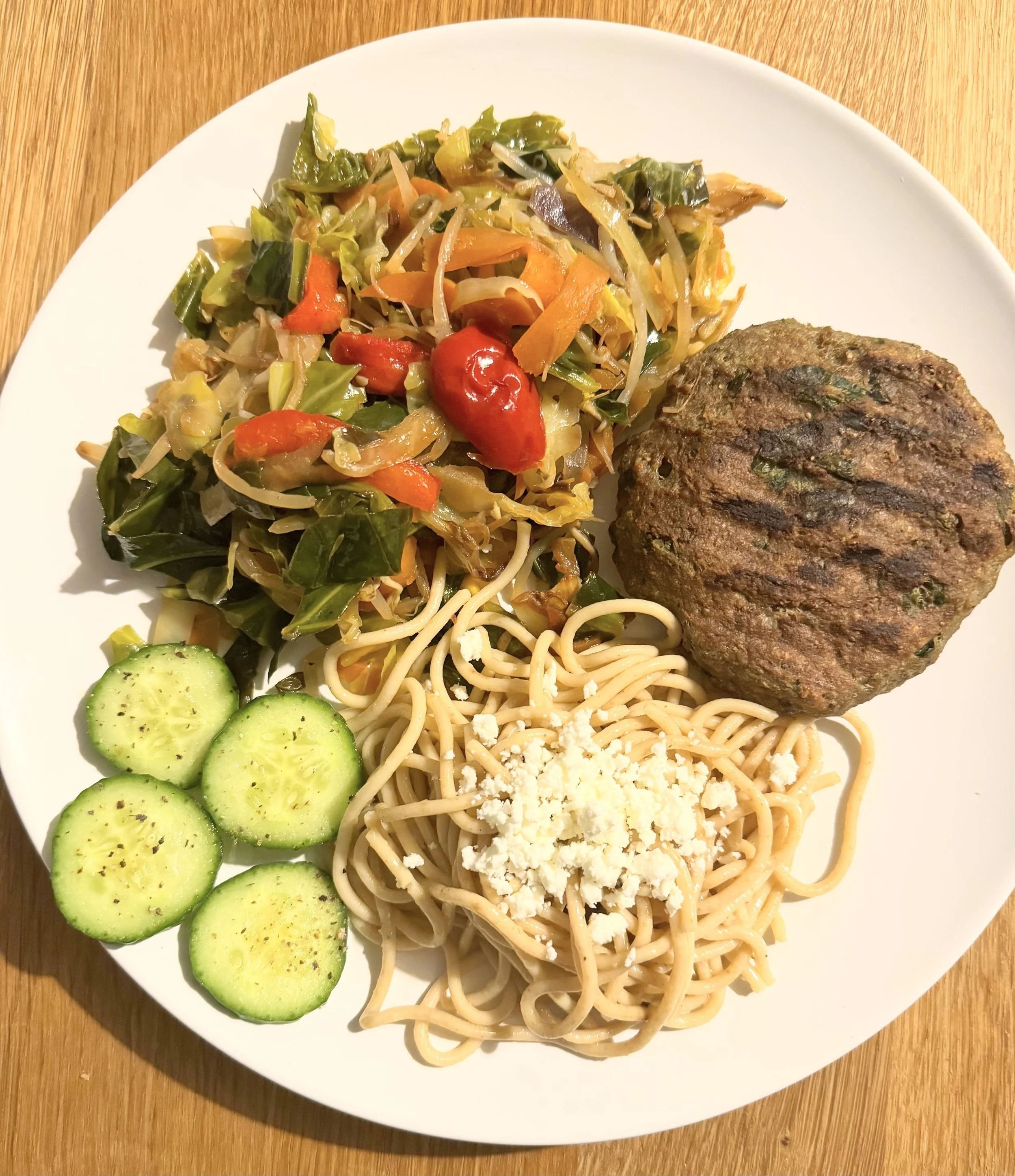Plate of food with stir-fried vegetables, grilled patty, spaghetti with cheese, and cucumber slices.