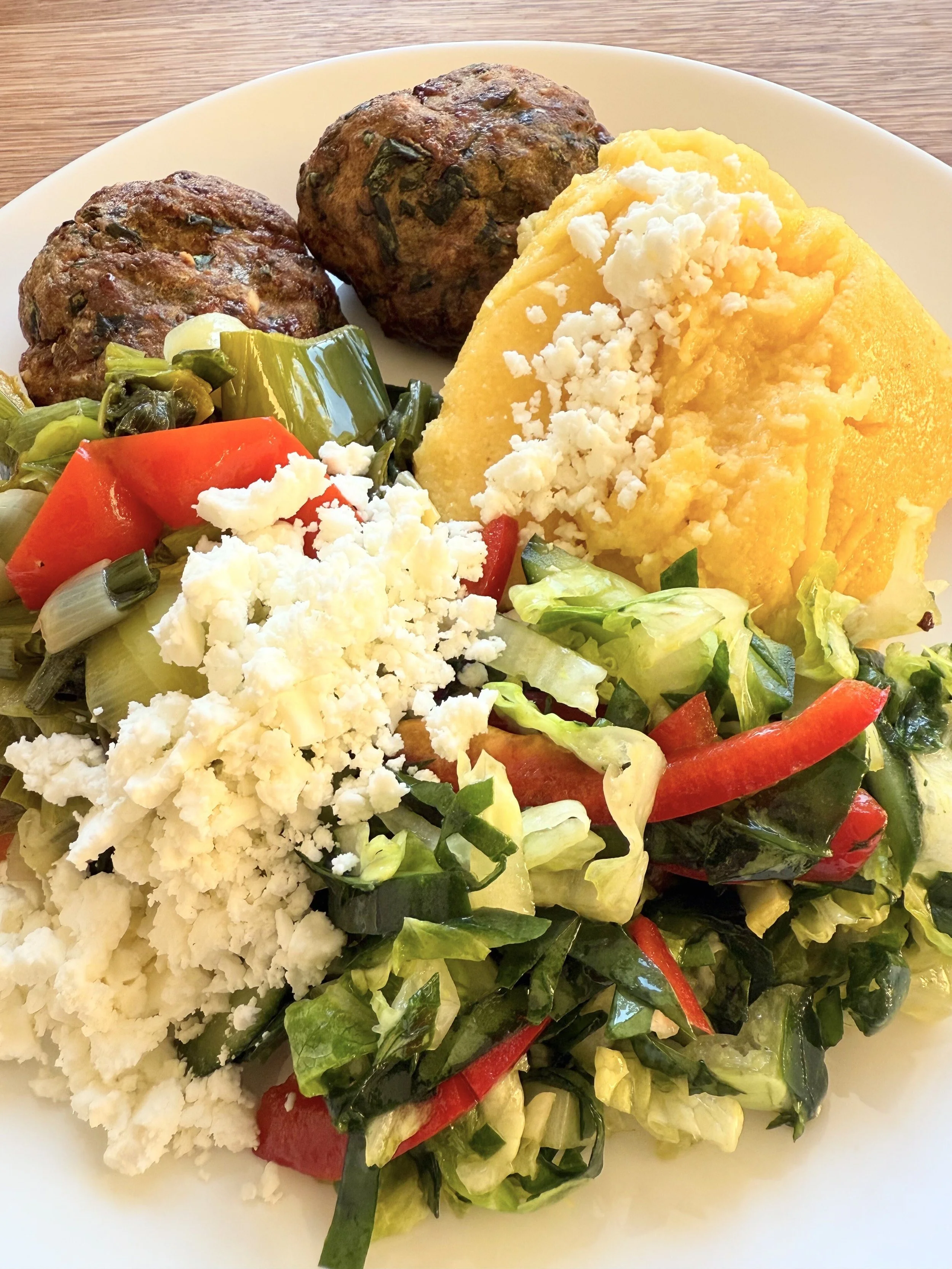 Plate with two meatballs, a scoop of yellow mashed potatoes topped with crumbled white cheese, and a side salad with mixed greens, chopped tomatoes, and shredded red peppers.
