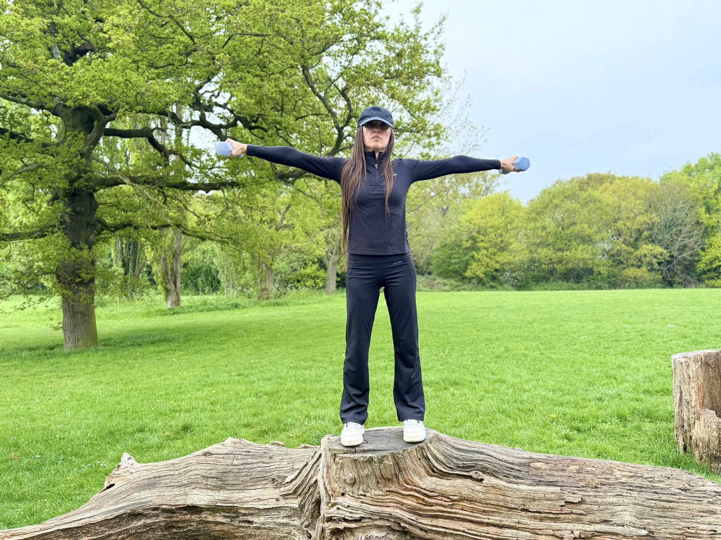 Woman standing on a fallen tree with arms outstretched, holding dumbbells, outdoors in a green park with trees and grass.